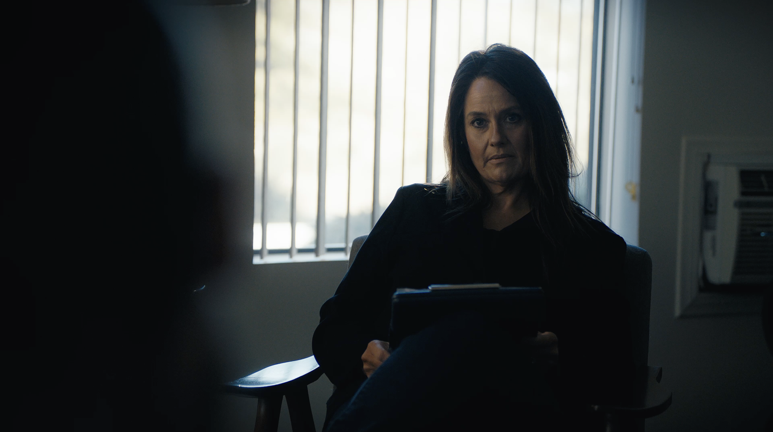 A woman with shoulder-length brown hair sitting in a dimly lit room, facing the camera, with a window with bars behind her. She appears serious or concerned, dressed in dark clothing, holding some documents or a folder on her lap.
