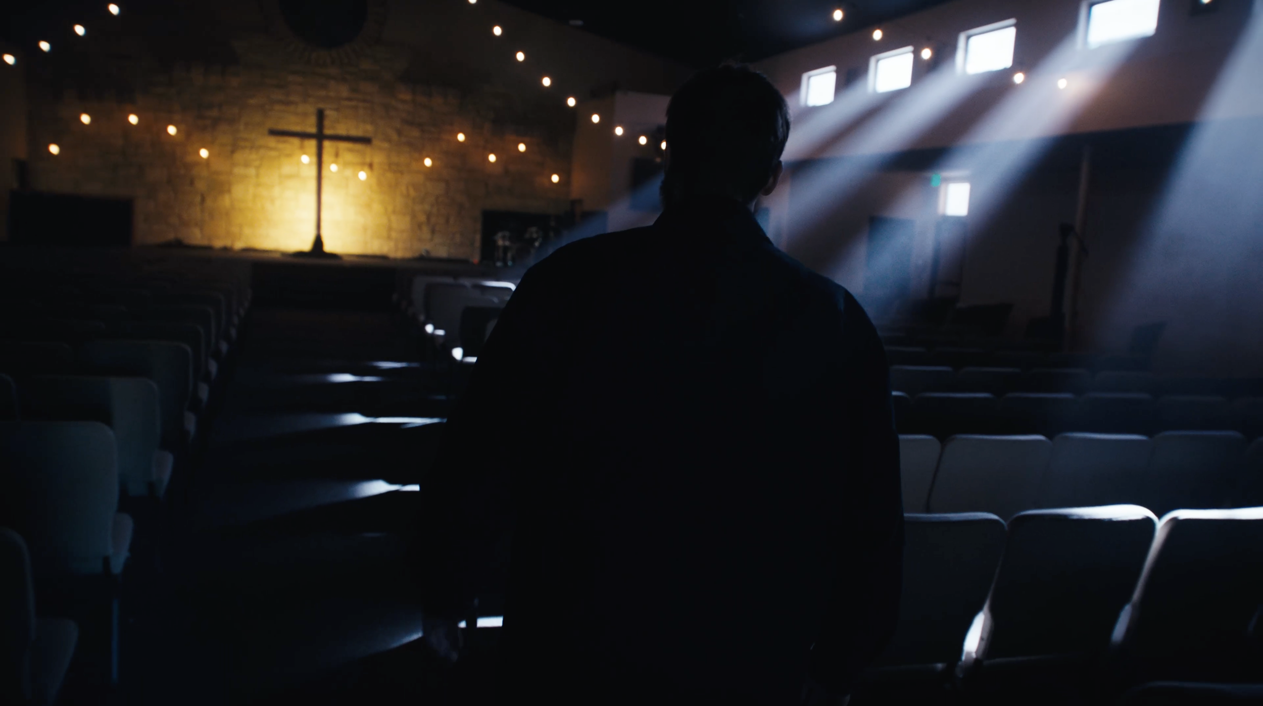 A man standing inside an empty church with a cross and brick wall at the front, dimly lit with warm lights and sunlight coming through windows.
