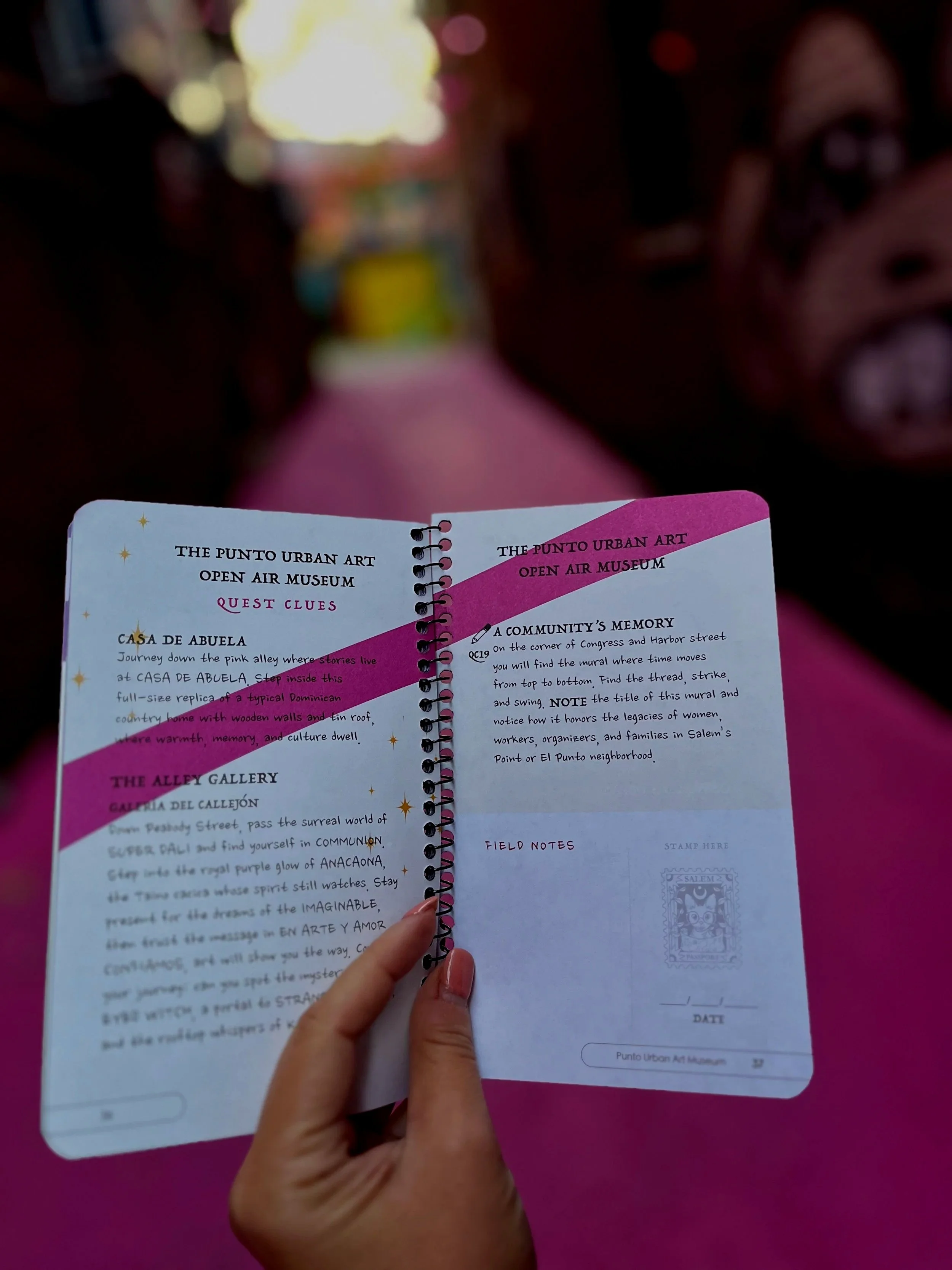 Open booklet with handwritten text and illustrations, held by a hand with pink nail polish, against a pink background.