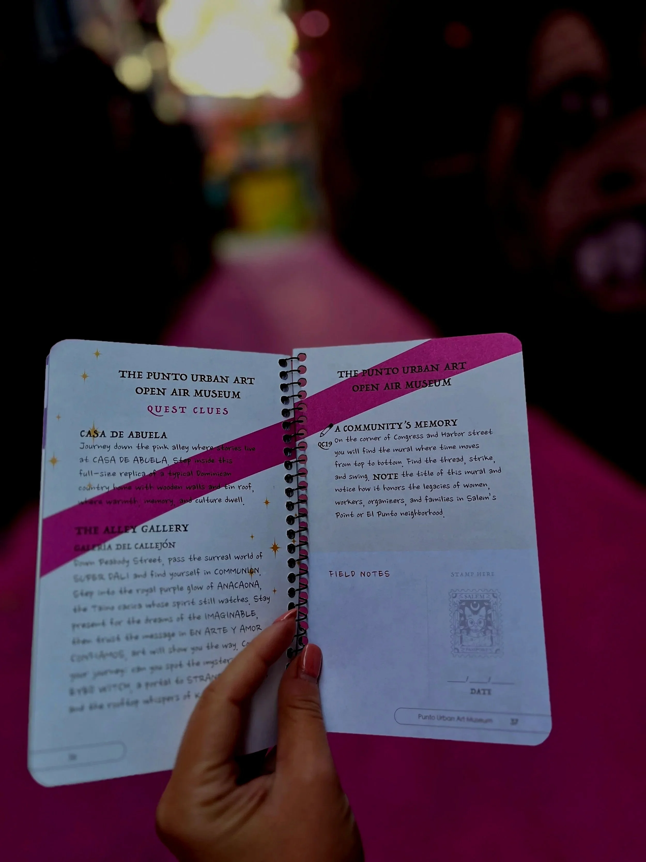 Open booklet with handwritten text and illustrations, held by a hand with pink nail polish, against a pink background.
