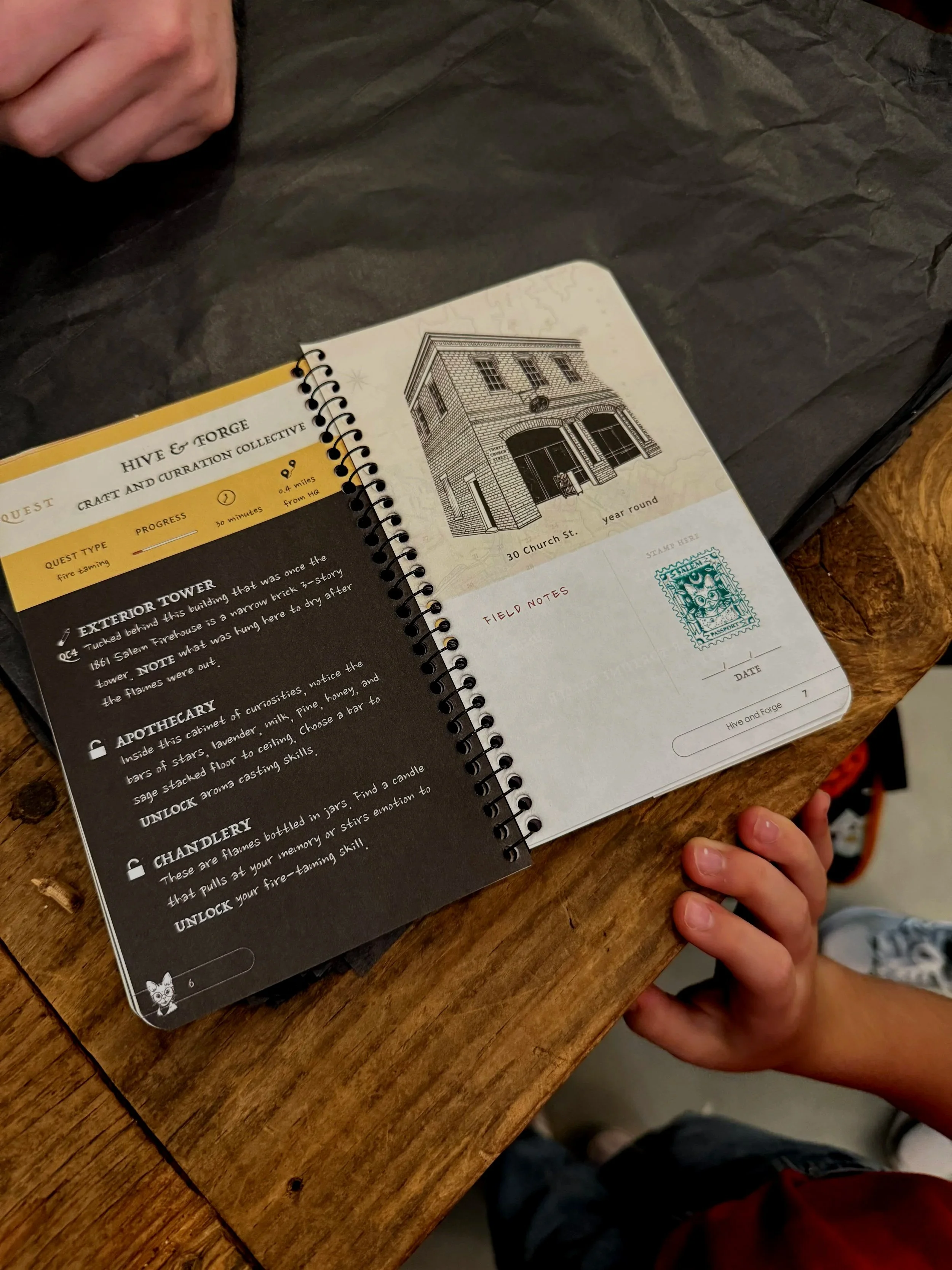 Close-up of an open illustrated journal on a wooden table, with a person's hand visible and a child's hand touching the edge of the table.