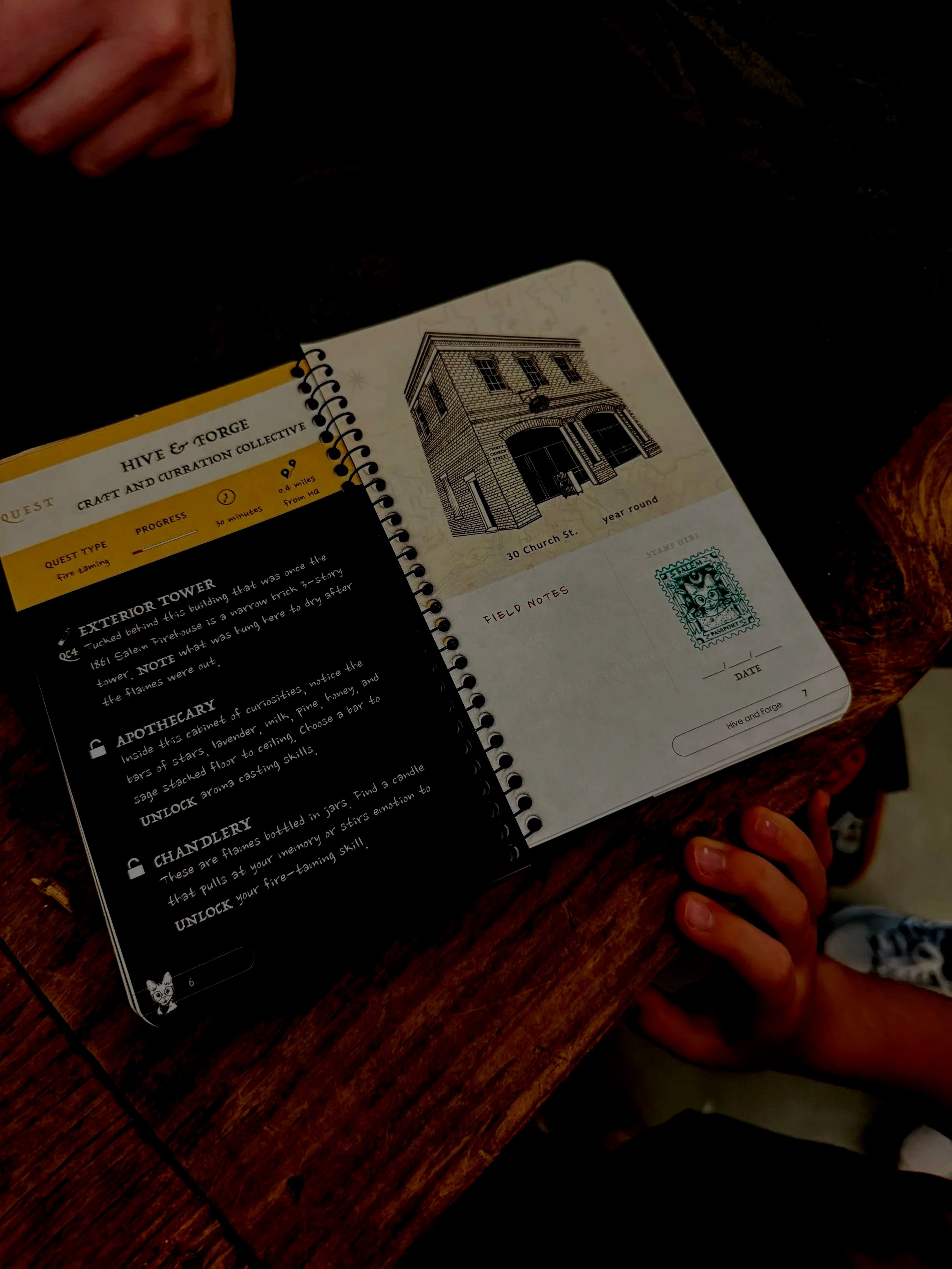 Close-up of an open illustrated journal on a wooden table, with a person's hand visible and a child's hand touching the edge of the table.