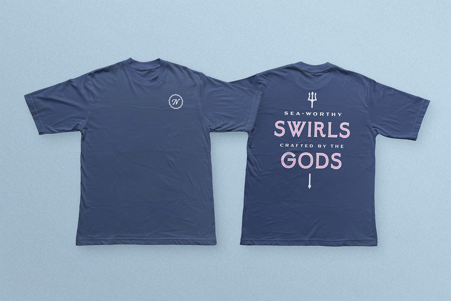 Two navy blue T-shirts laid flat on a light blue background. The front of the shirt features a small white logo on the left chest, and the back has larger text reading 'SEA-WORTHY SWIRLS CRAFTED BY THE GODS,' with a small trident graphic above the te