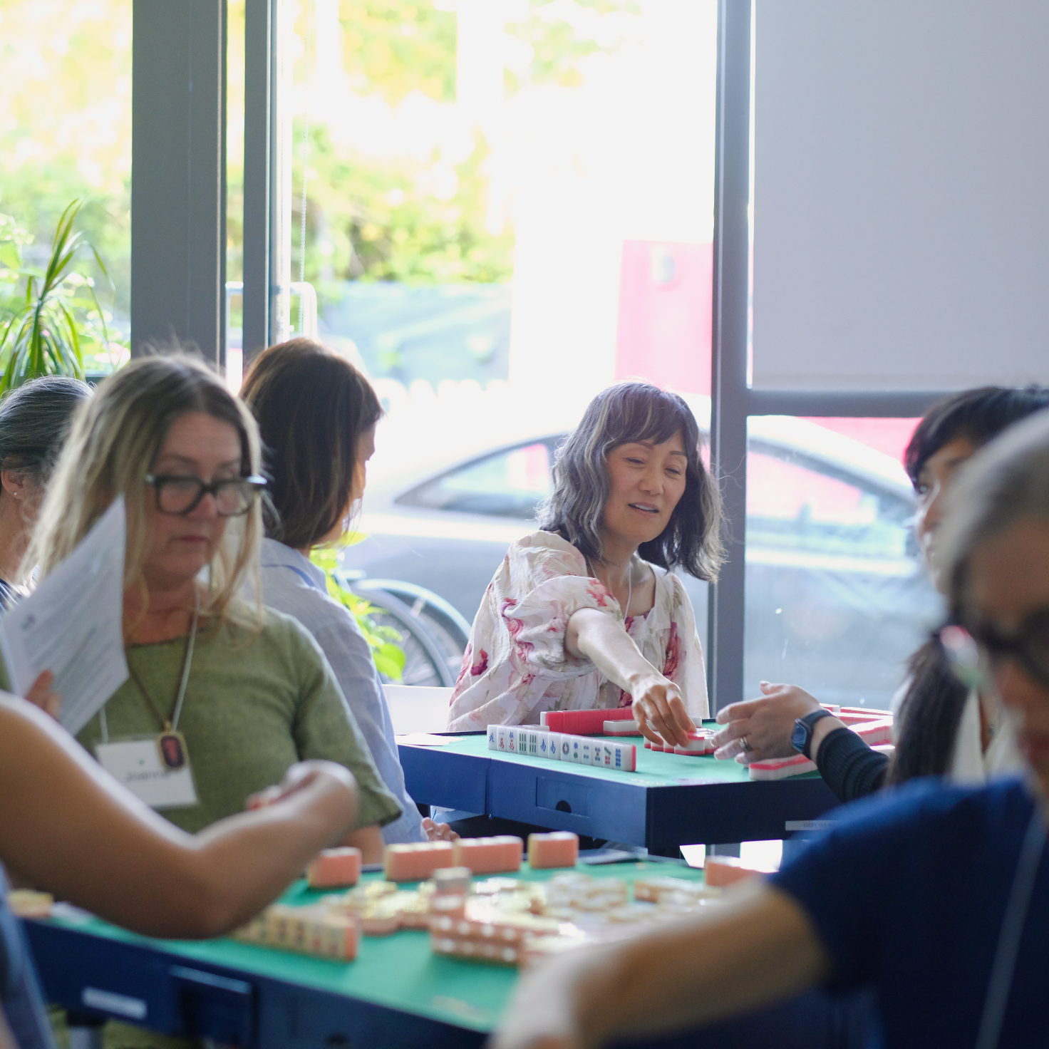 People playing Mahjong at a daytime gathering inside a bright room near large windows.