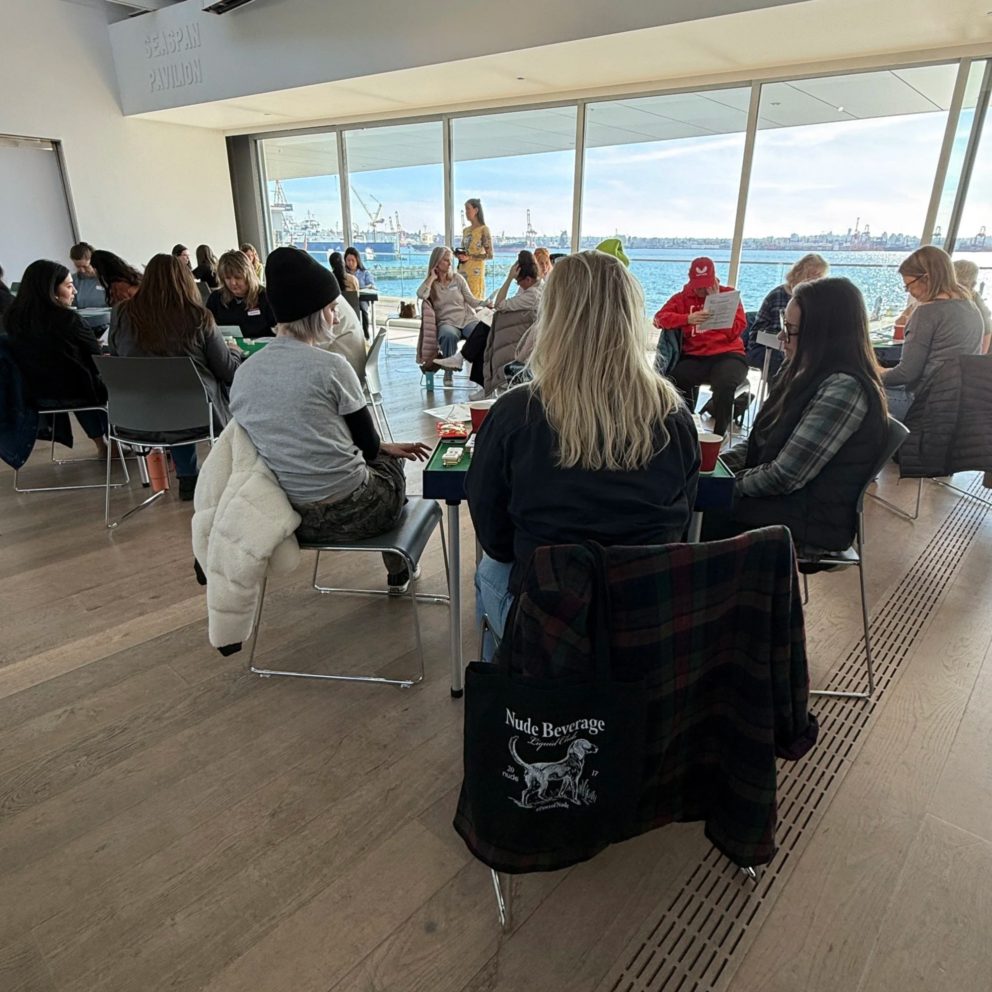 A group of people sitting at mahjong tables in a bright indoor space with large windows overlooking a body of water.