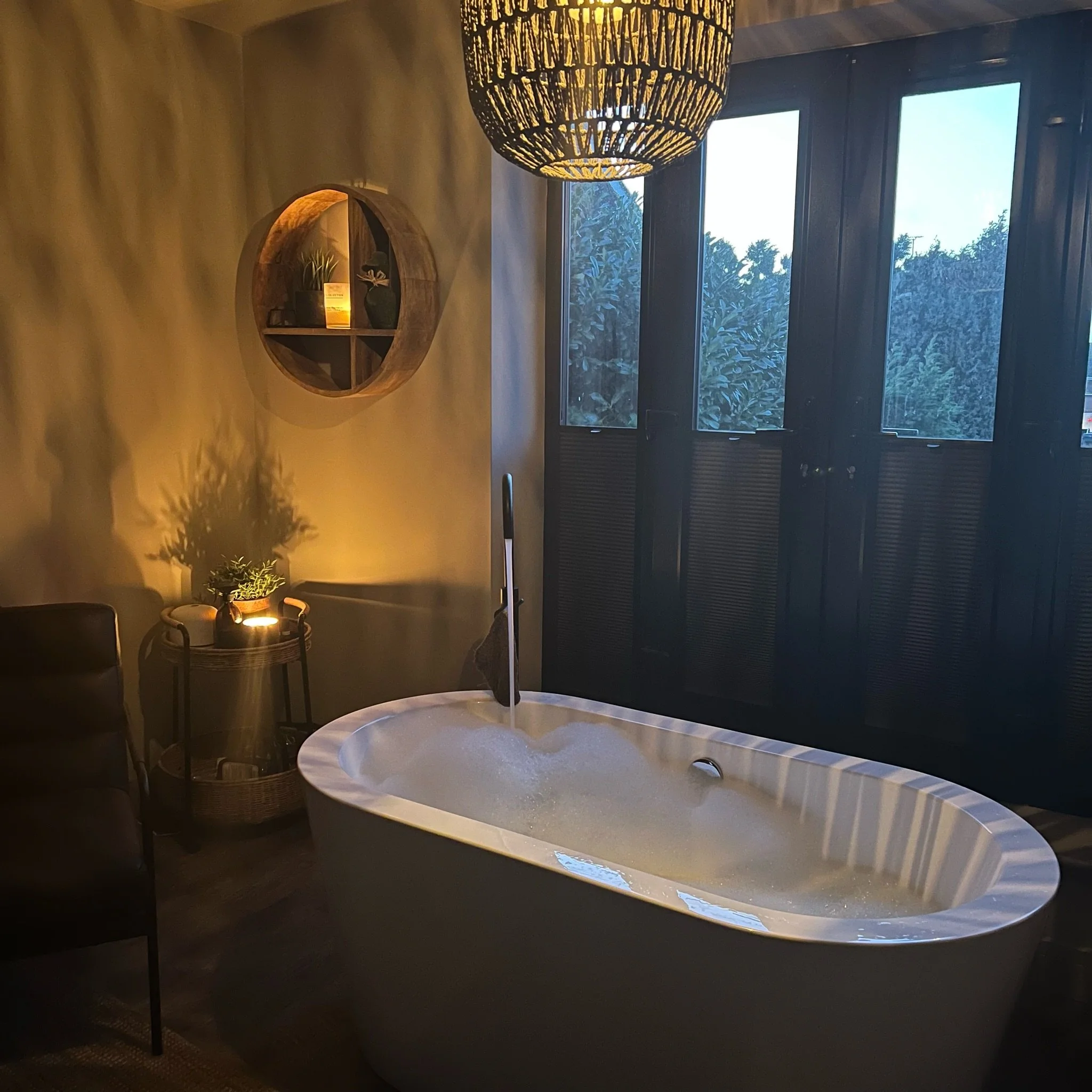 A bathroom in Gloucestershire with a large white bathtub, a wooden circular wall shelf with decorative objects, a small table with a plant and candle, and a window with dark shutters. A light hangs from the ceiling, casting shadows on the wall.