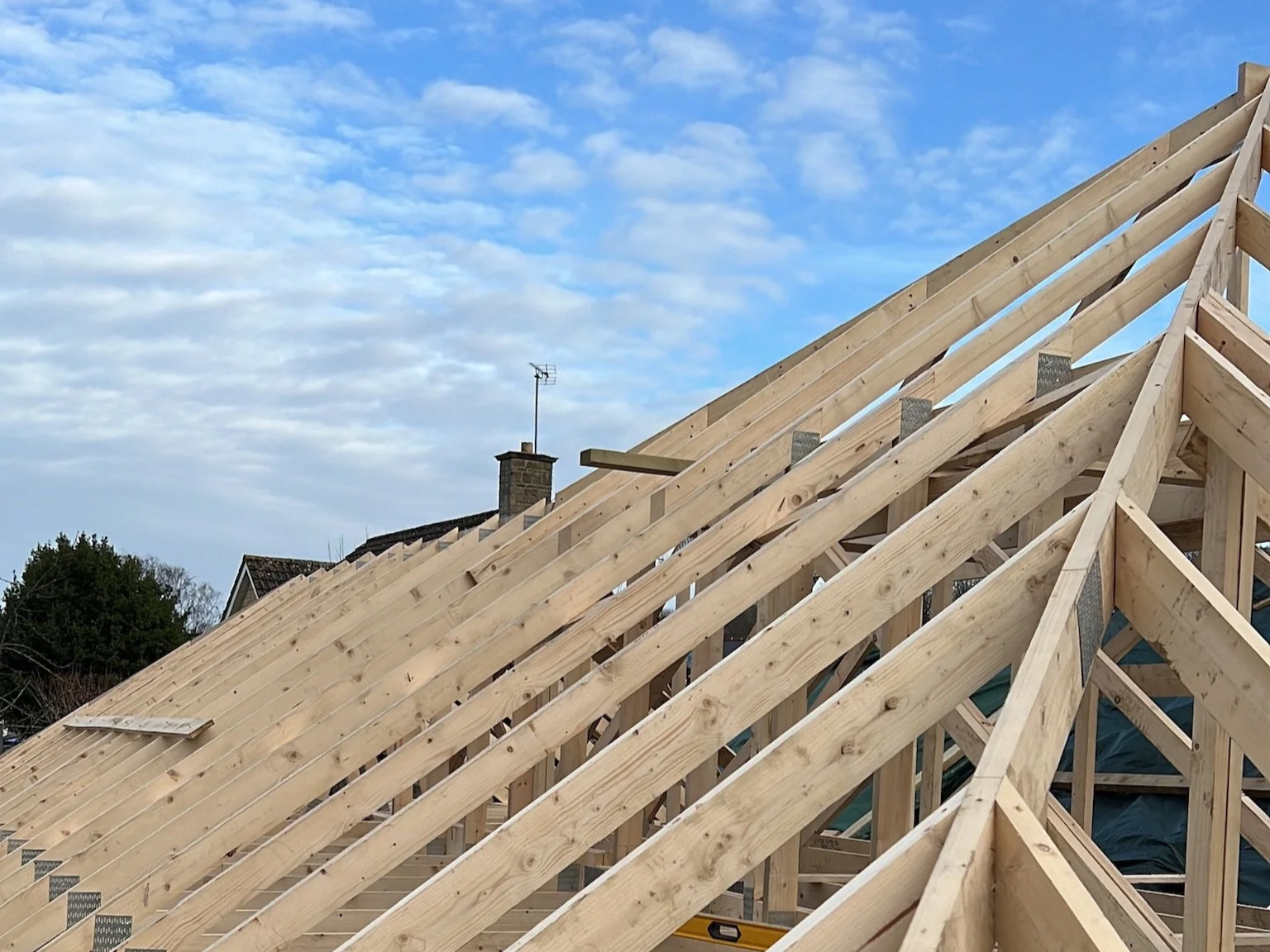 Construction site in The Cotswolds with a wooden roof frame in progress, under a blue sky with scattered clouds.