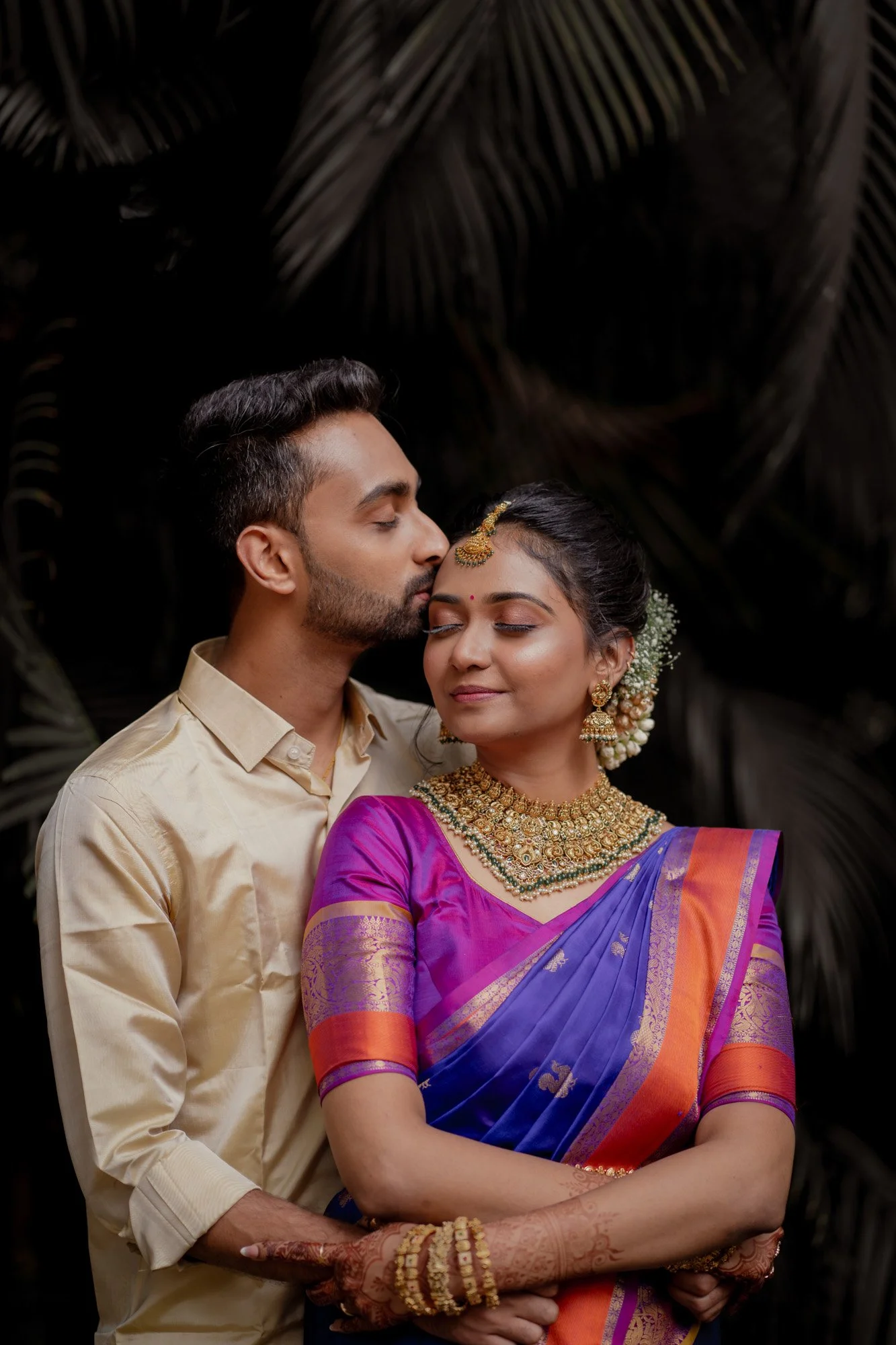 Couple in traditional attire, woman in a blue and pink sari with gold jewelry, man in a beige shirt, embracing.