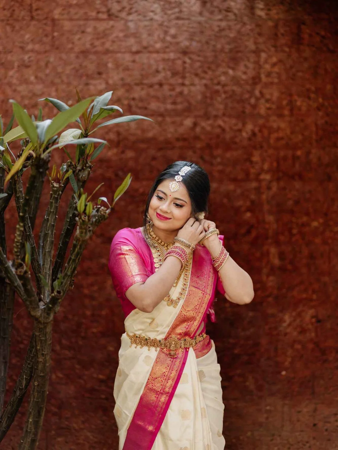 Woman in traditional Indian attire adjusting her earring, wearing a white and pink sari, with gold jewelry, standing next to a small plant against a brown textured background.