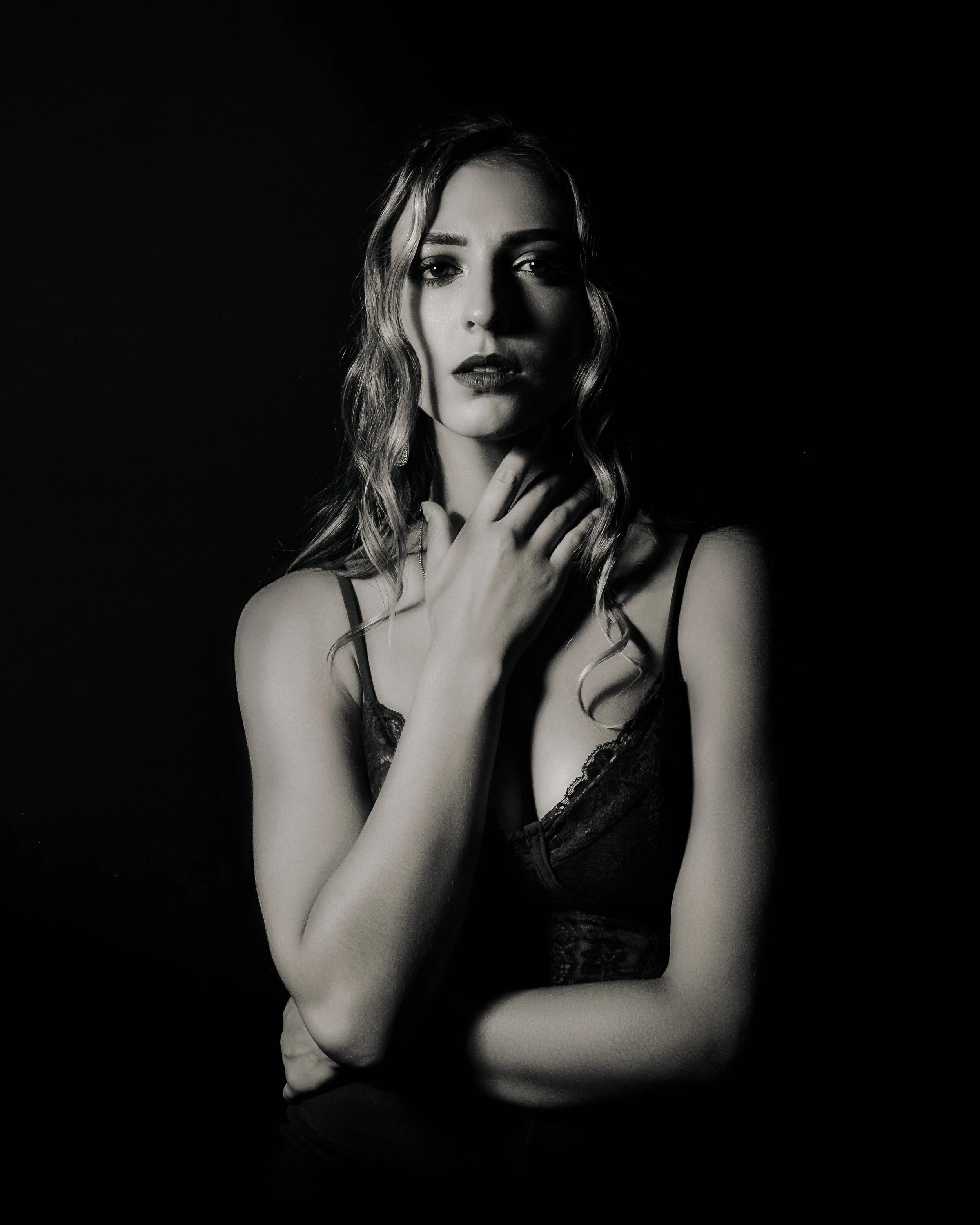 A black and white portrait of a woman with wavy hair, wearing a lace top, posing with her hand near her neck against a dark background.