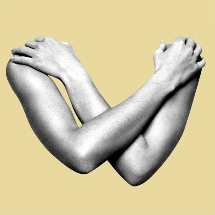 Black and white photo of two human arms crossed to form a heart shape, set against a beige background.