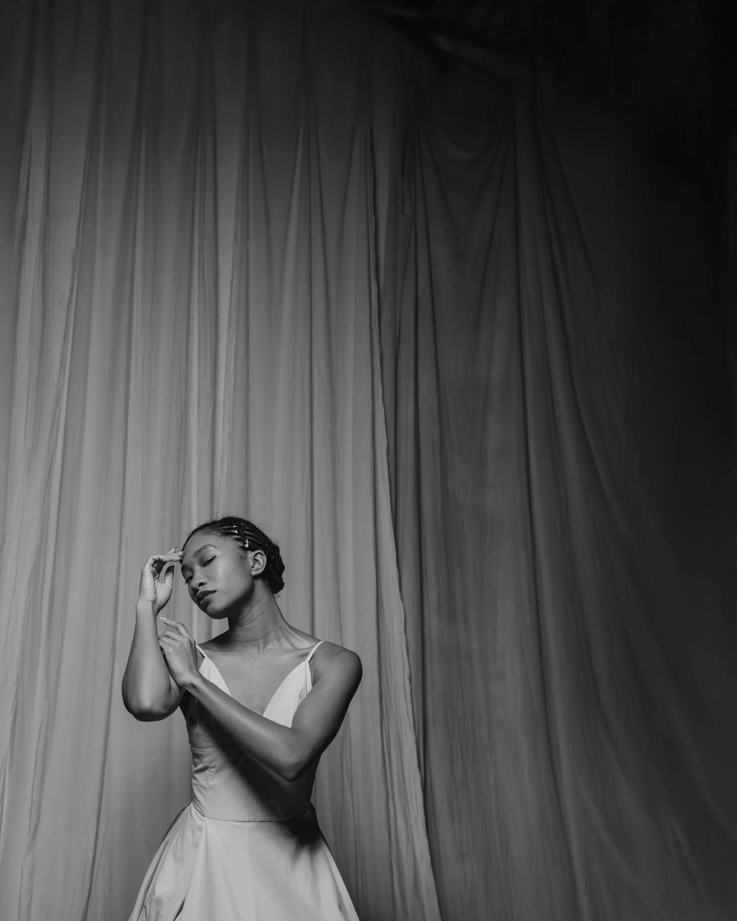 A woman in a light-colored dress stands in front of a curtain, with her eyes closed and hand touching her forehead, appearing contemplative.