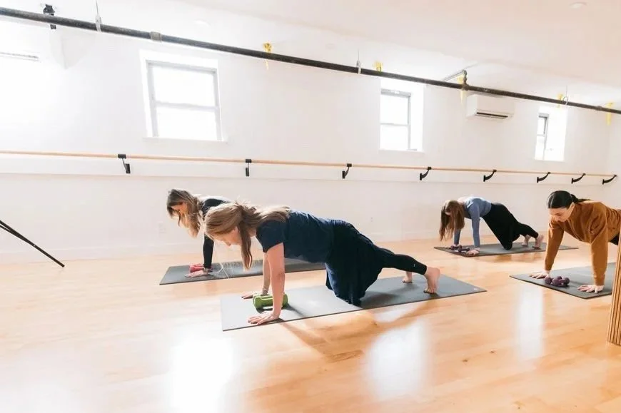 Two women exercising in a gym, one lifting a pink dumbbell above her head and the other holding a pink dumbbell while balancing on one leg.
