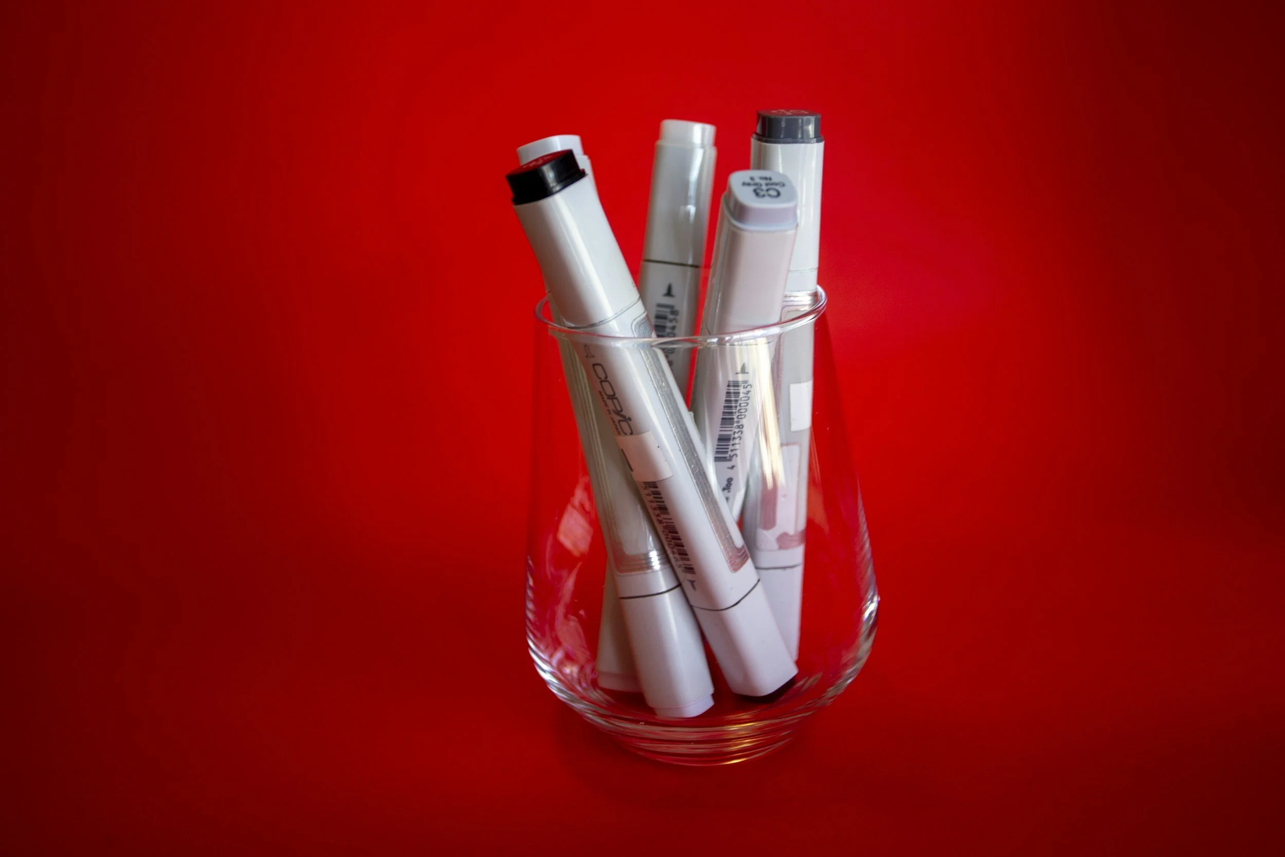 A clear glass holding white markers with black and gray caps on a red background.
