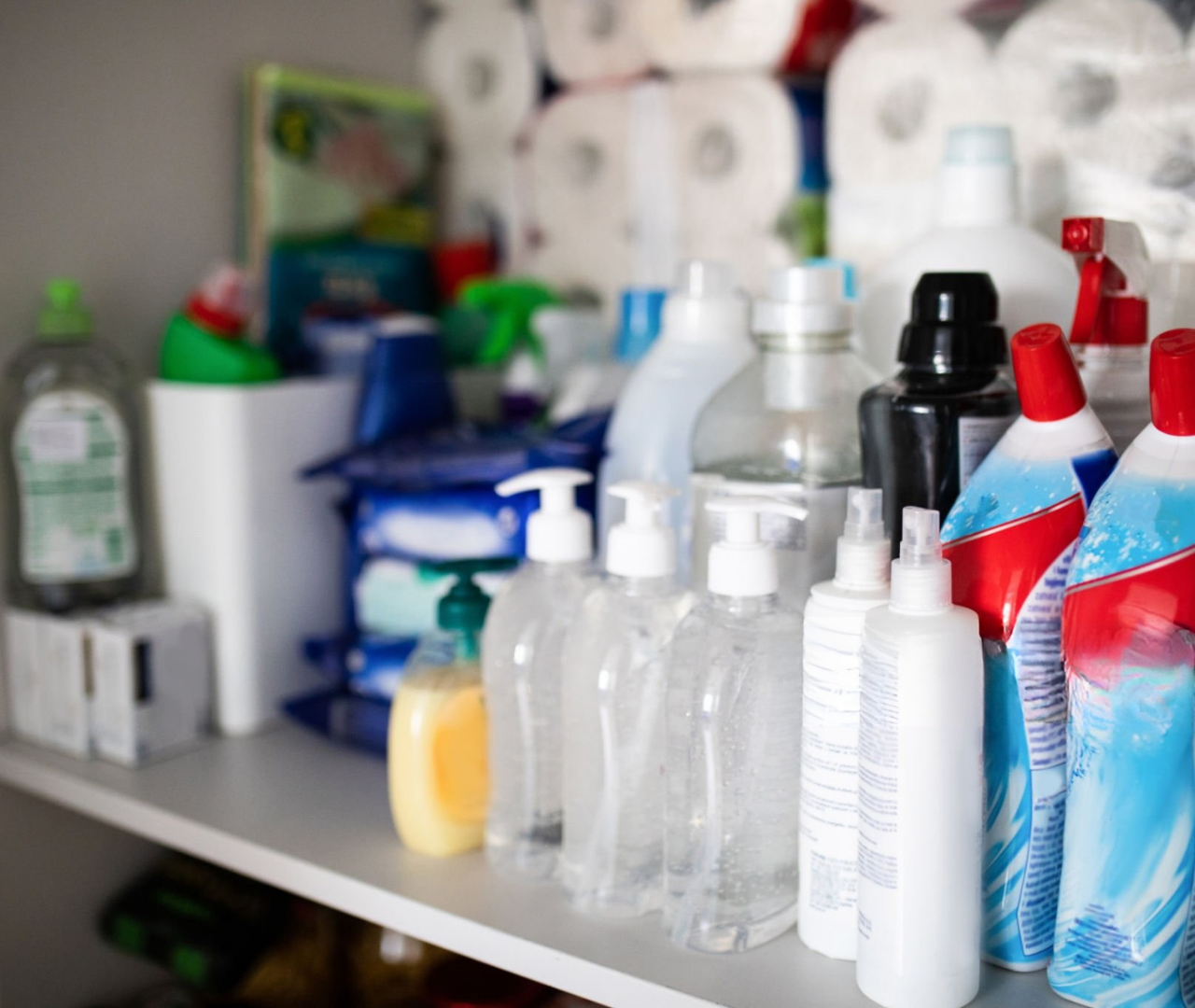 Shelf filled with various household cleaning supplies, including spray bottles, liquid soap dispensers, and boxes of toilet paper.