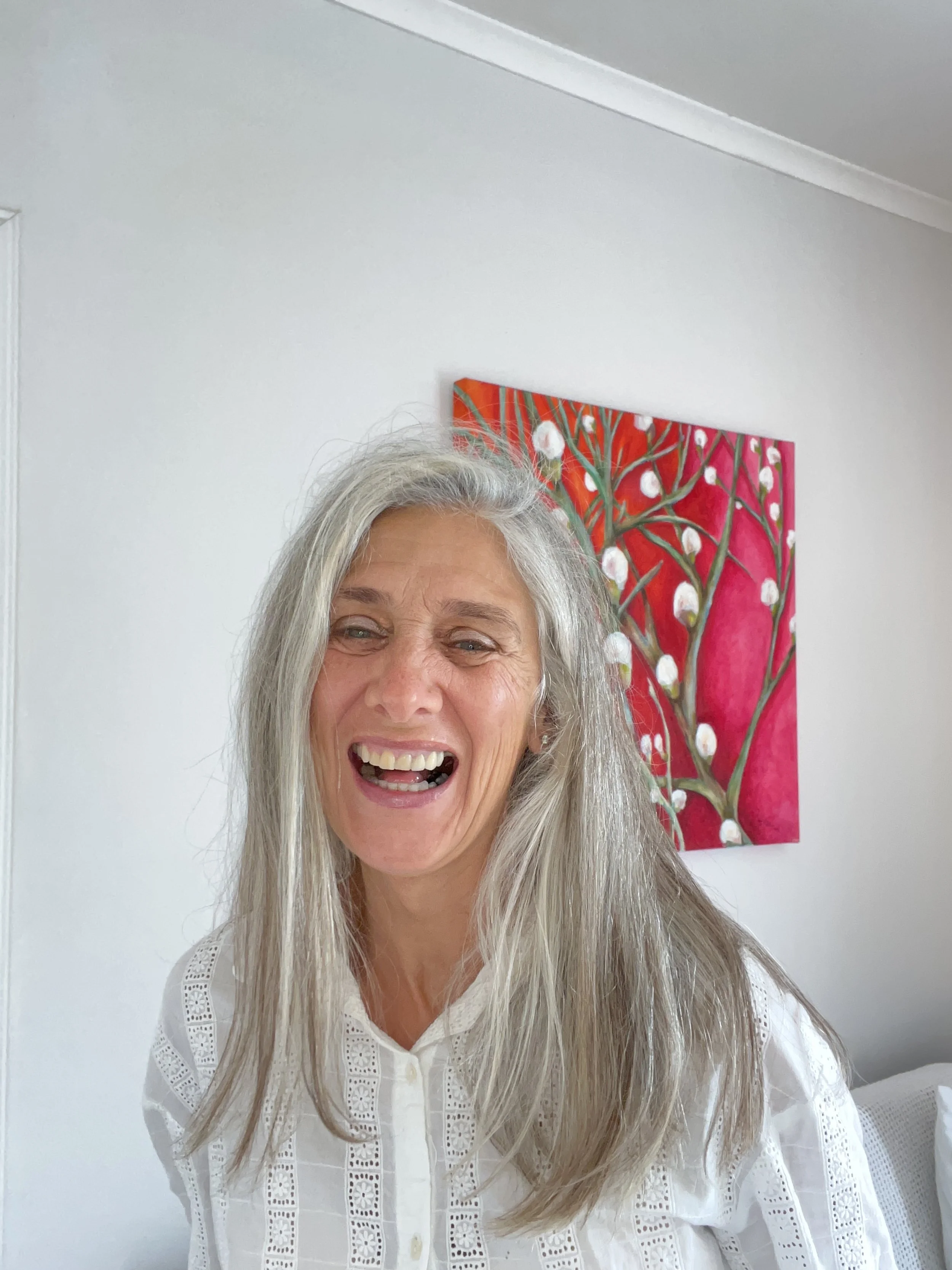 A smiling older woman with long gray hair in a white blouse, standing in front of a white wall with colorful abstract floral artwork.