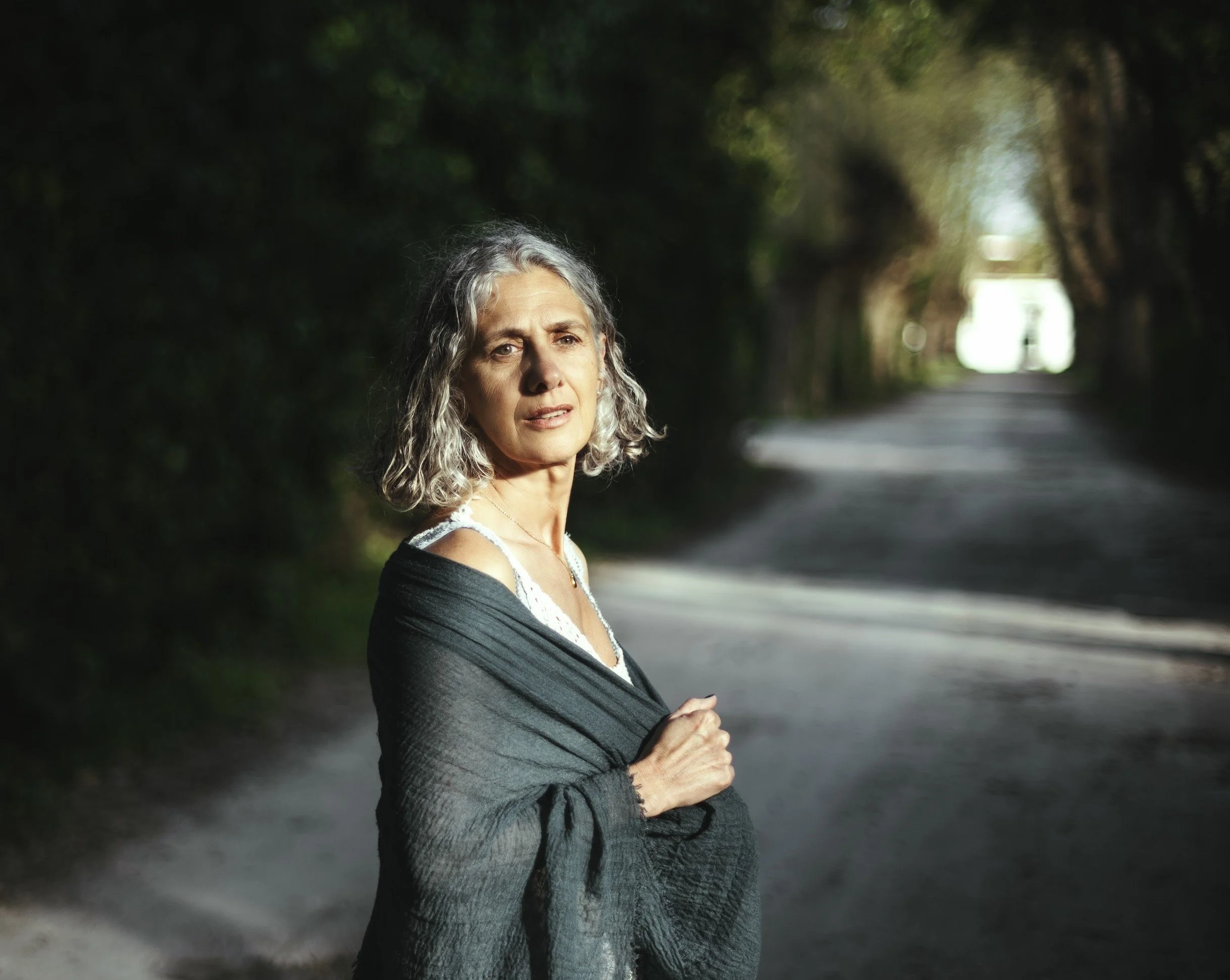 A woman with curly gray hair stands on a tree-lined road, looking into the distance with a concerned or thoughtful expression, holding a dark shawl over her shoulders.