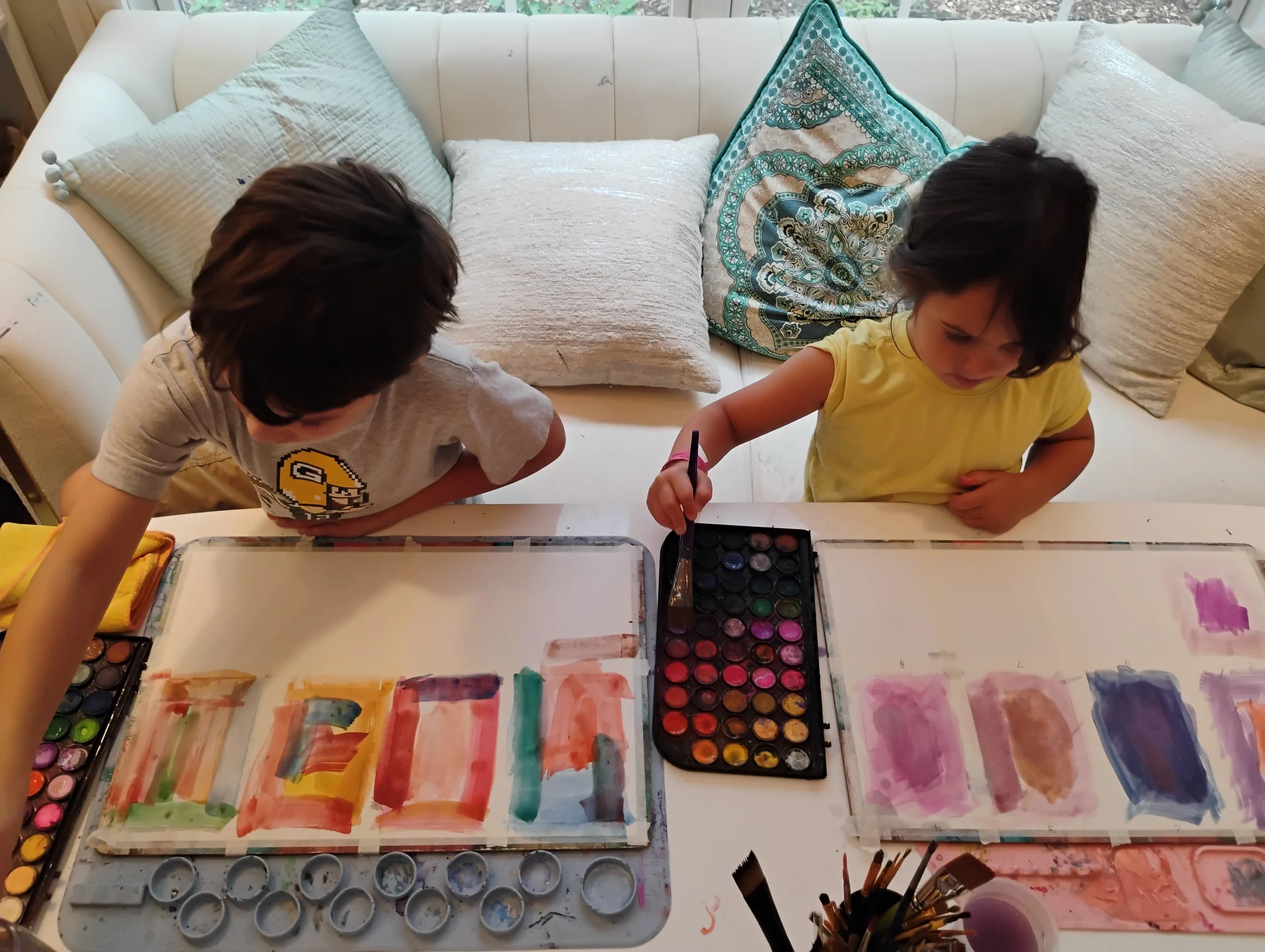 Two children sit at a table with watercolor paints, creating artwork at Lunisa summer camp. The table is covered with rainbow-colored watercolor paintings, and there are paint palettes and brushes visible.