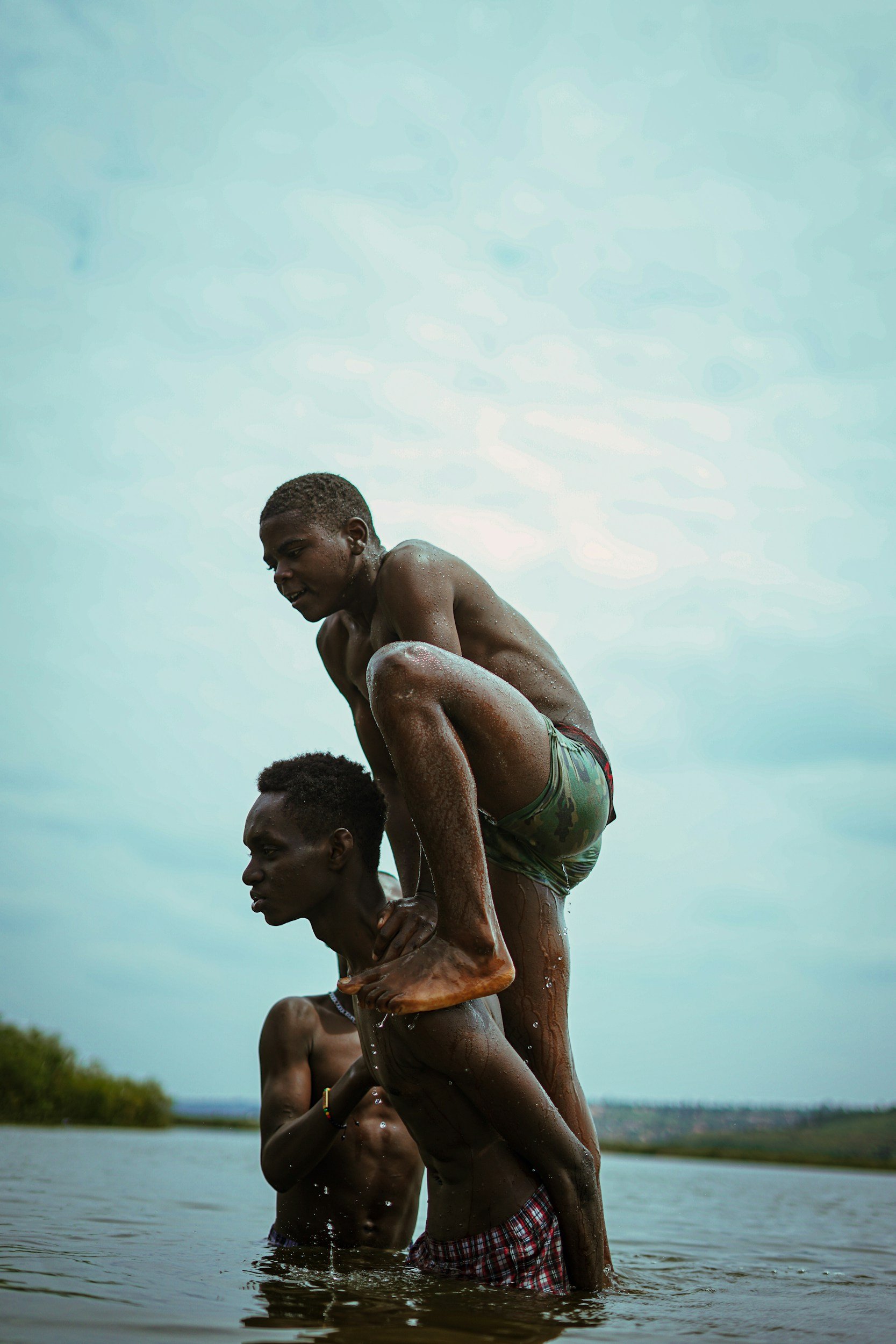 Two young men are balancing on each other's shoulders in a body of water, with one standing on the other's shoulders. The scene appears to be outdoors with a cloudy sky and a distant landscape visible in the background.