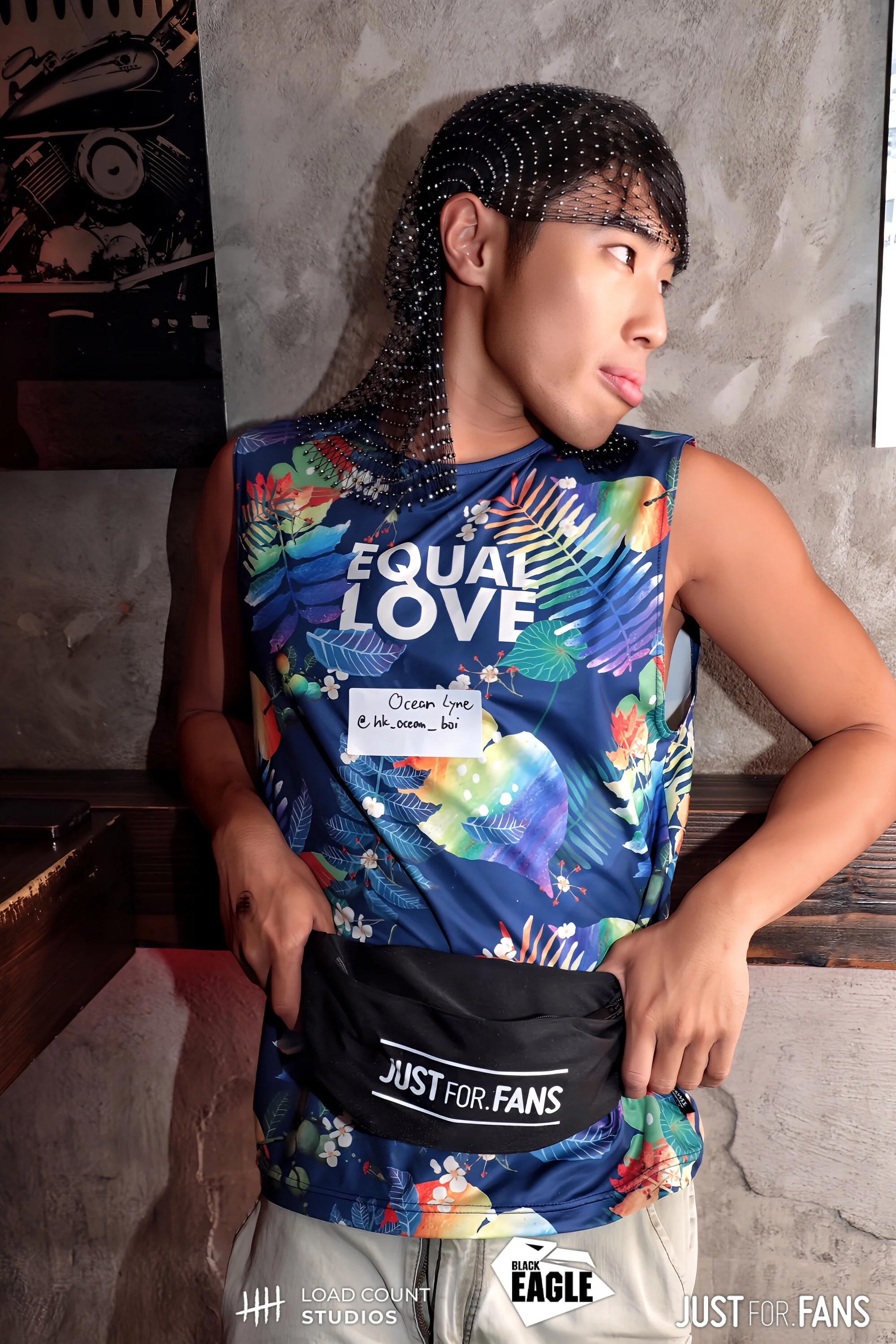 Person wearing a colorful sleeveless top with 'EQUAL LOVE' text, a mesh headpiece, and holding a 'JUST FOR FANS' bag in an indoor setting.