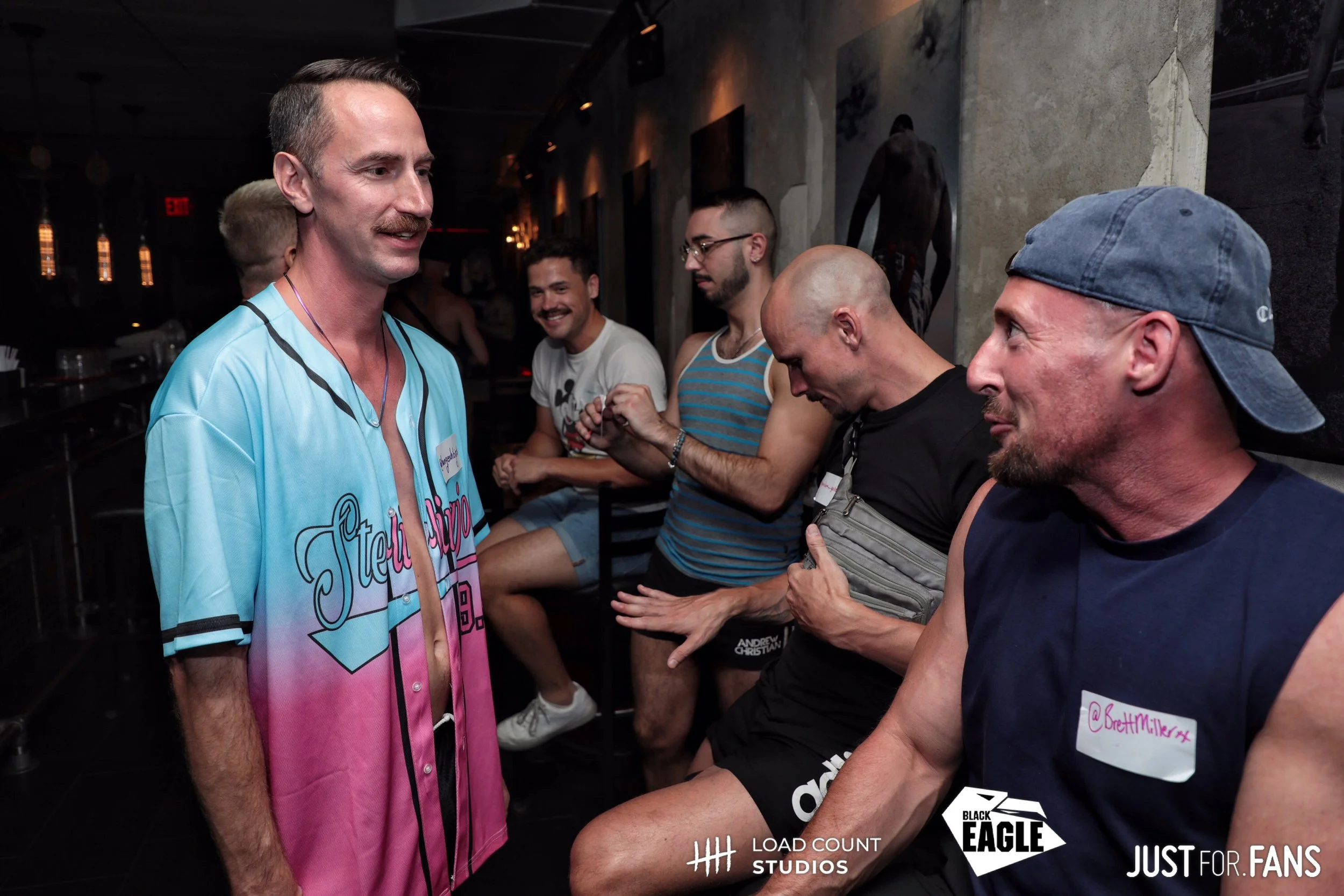 Group of men in casual attire at a bar, some wearing name tags, engaging in conversation and laughter. Dimly lit, with background art and branding logos visible.