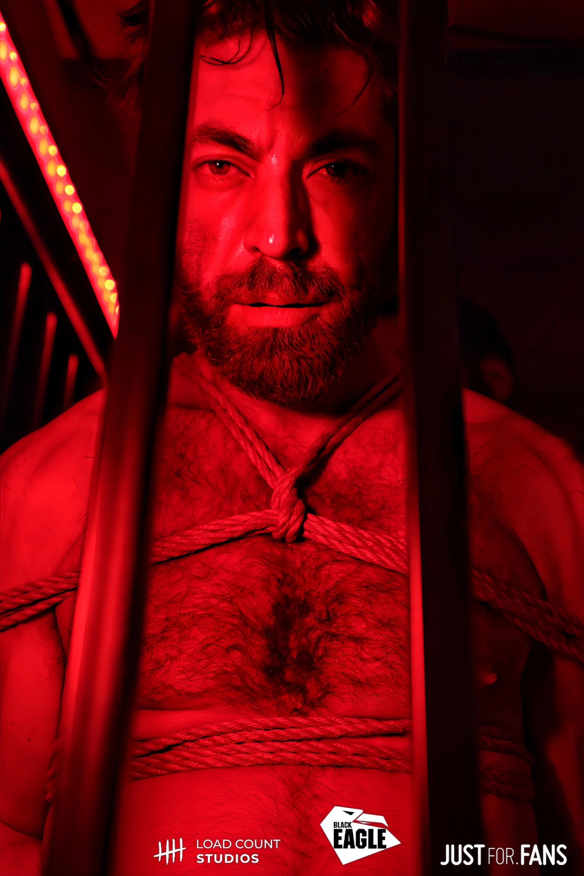 Man with beard behind bars, tied with ropes, in red lighting