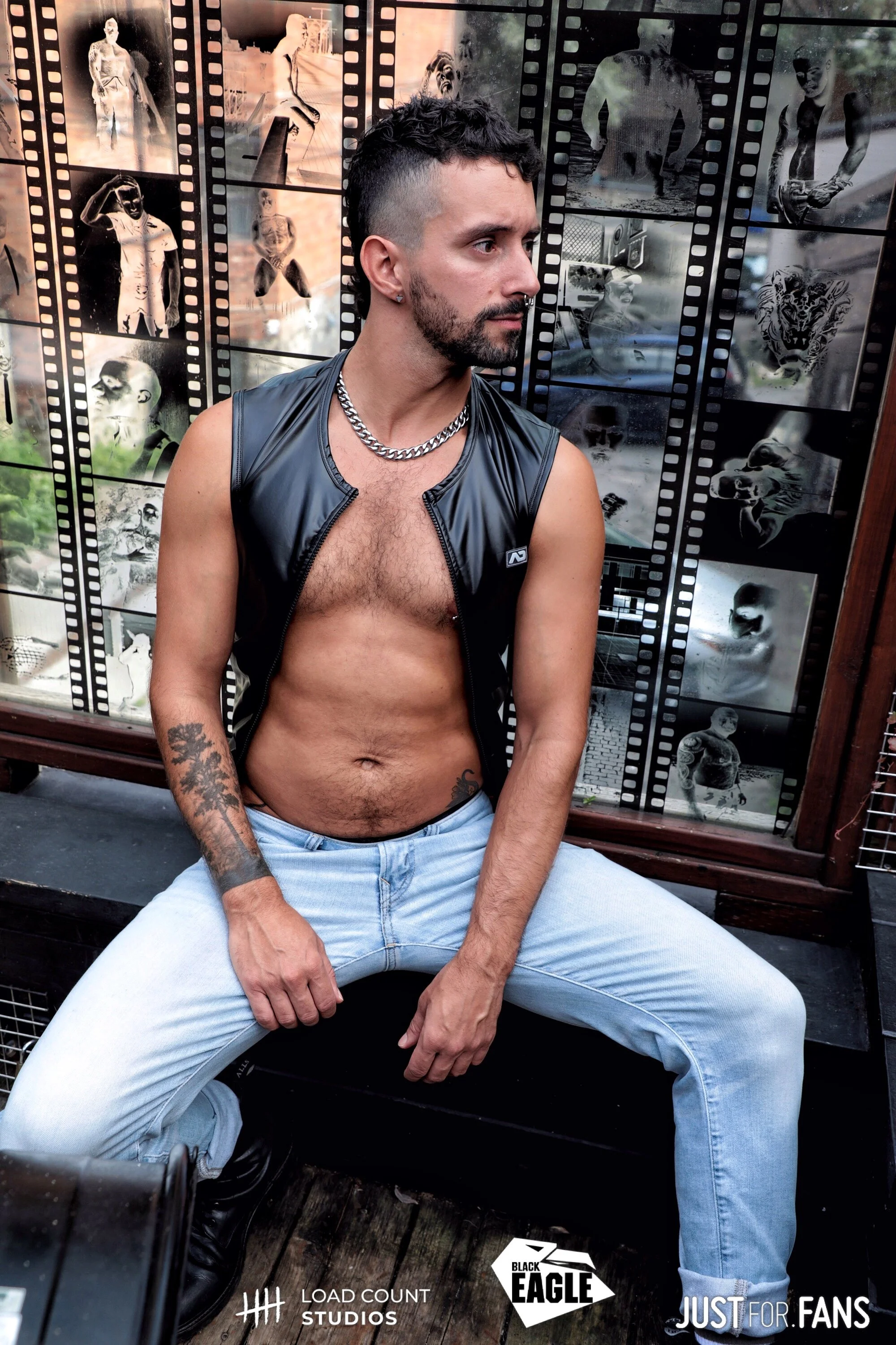 Man in open leather vest and jeans sitting in front of a wall covered with film strip images.