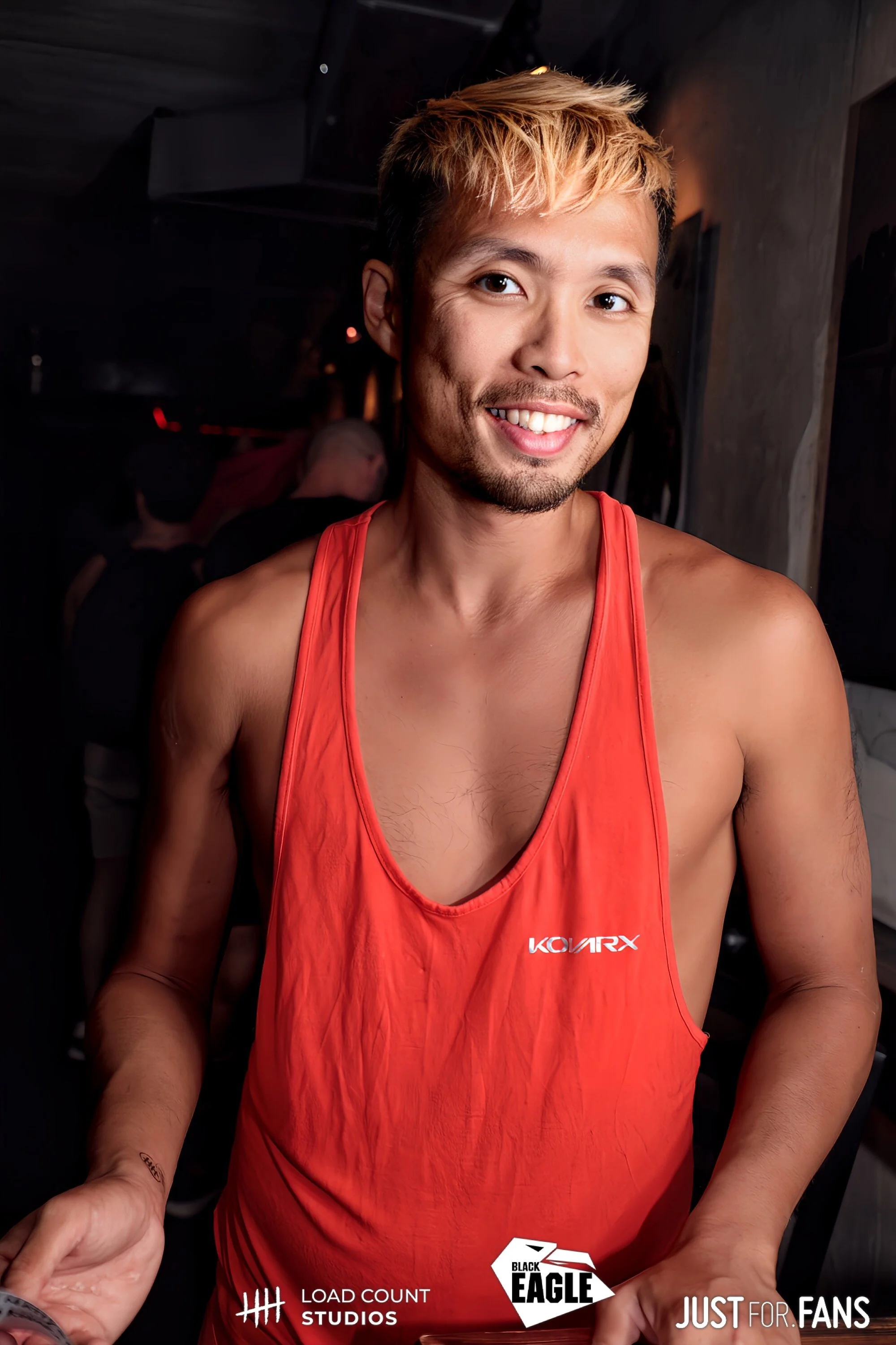 Person in a red tank top at a dimly lit venue with a logo on the shirt, smiling at the camera.