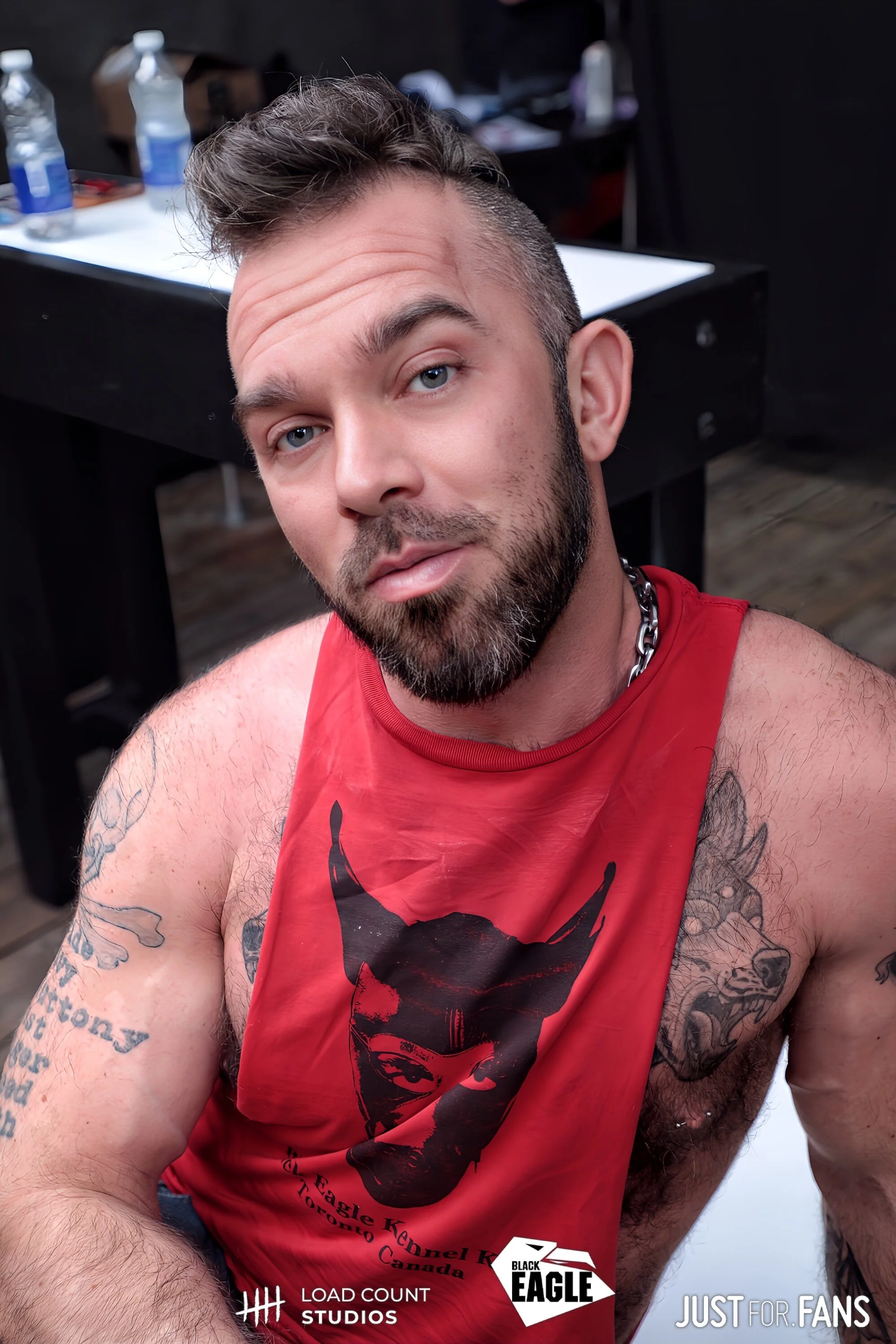 Man with styled hair, beard, wearing a red tank top with graphic design, seated in front of a table with water bottles in the background.