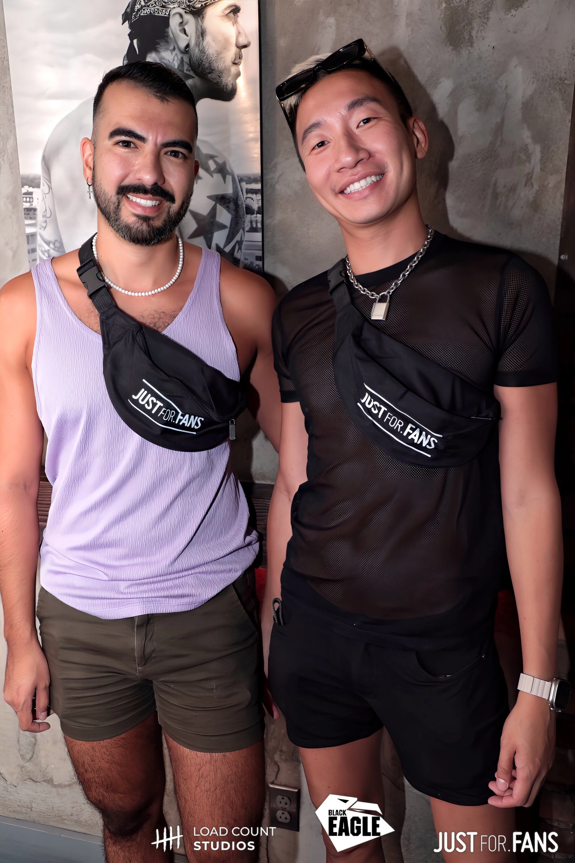 Two people posing for a photo inside a venue, both wearing "Just For Fans" branded bags. One is in a light purple tank top and shorts, the other in a black mesh shirt. Logos for Load Count Studios, Black Eagle, and Just For Fans are visible at the bo