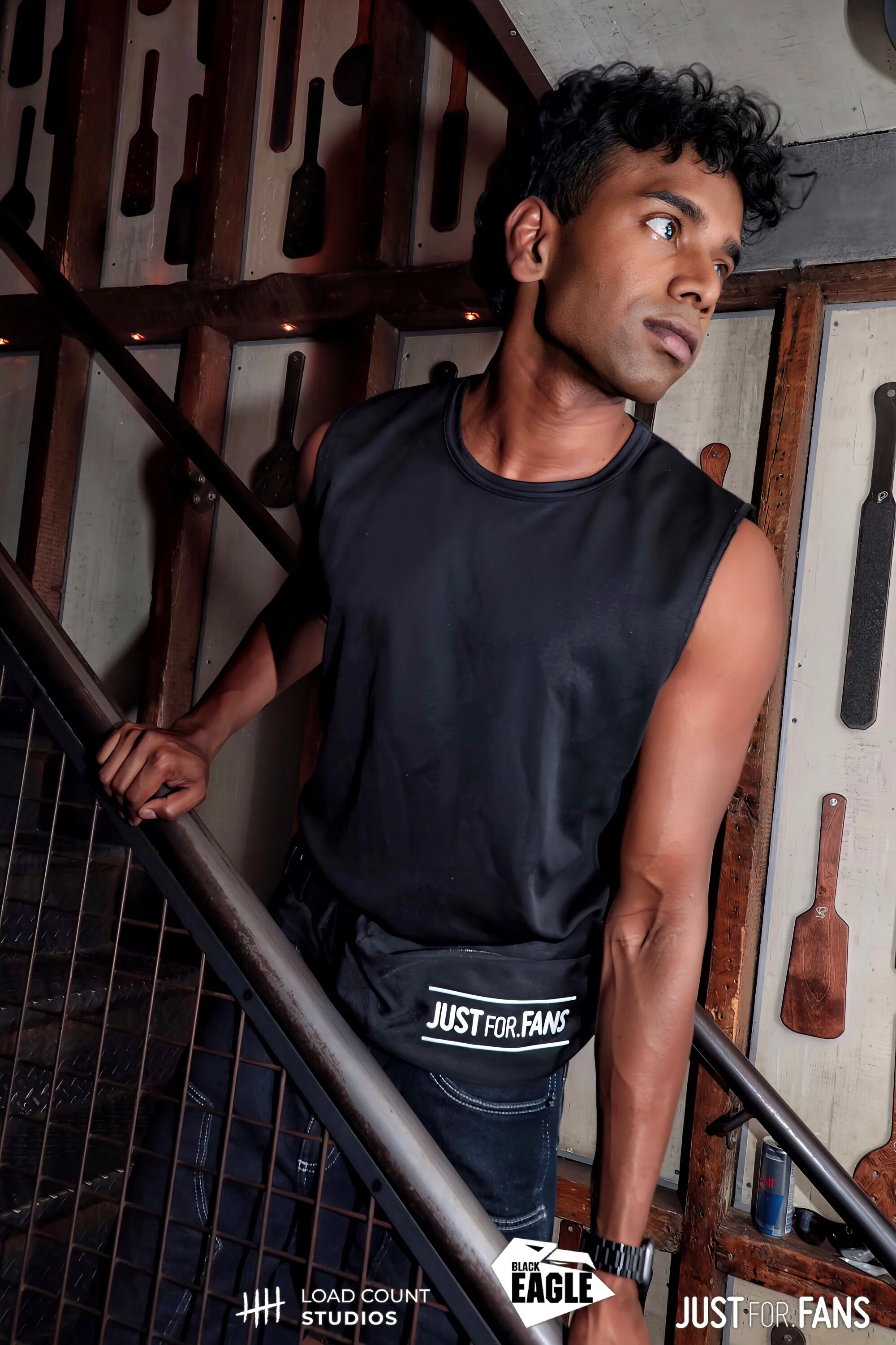 Person leaning against a metal stair railing, dressed in a black sleeveless shirt and wearing a fanny pack with 'Just For Fans' branding, surrounded by a wall with hanging paddles and logos for Load Count Studios, Black Eagle, and Just For Fans.