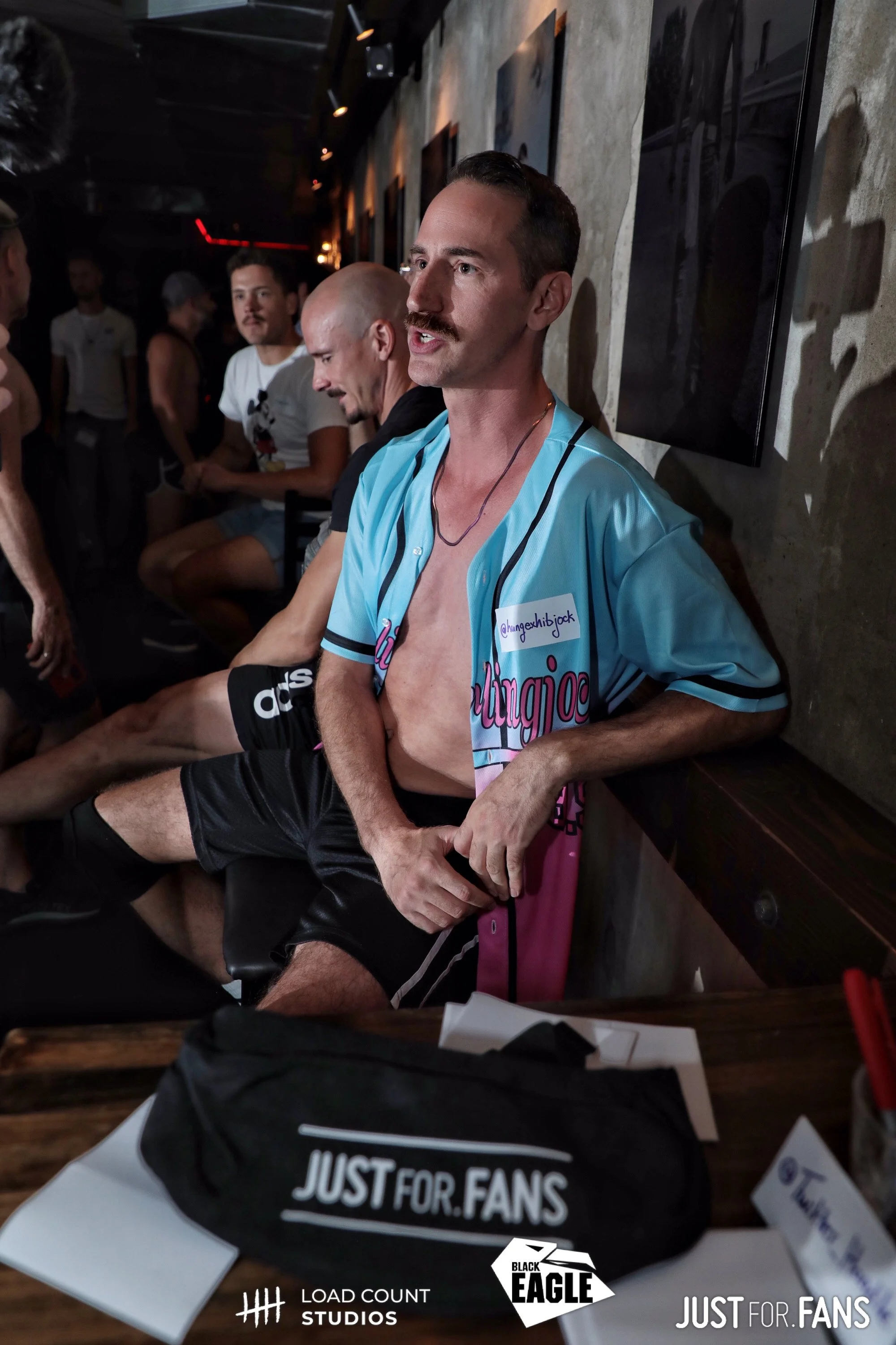 A group of men in a dimly lit room, with one man in the foreground wearing a blue baseball jersey. The setting appears to be a social event, with promotional bags labeled "Just For Fans" and logos for "Load Count Studios" and "Black Eagle."