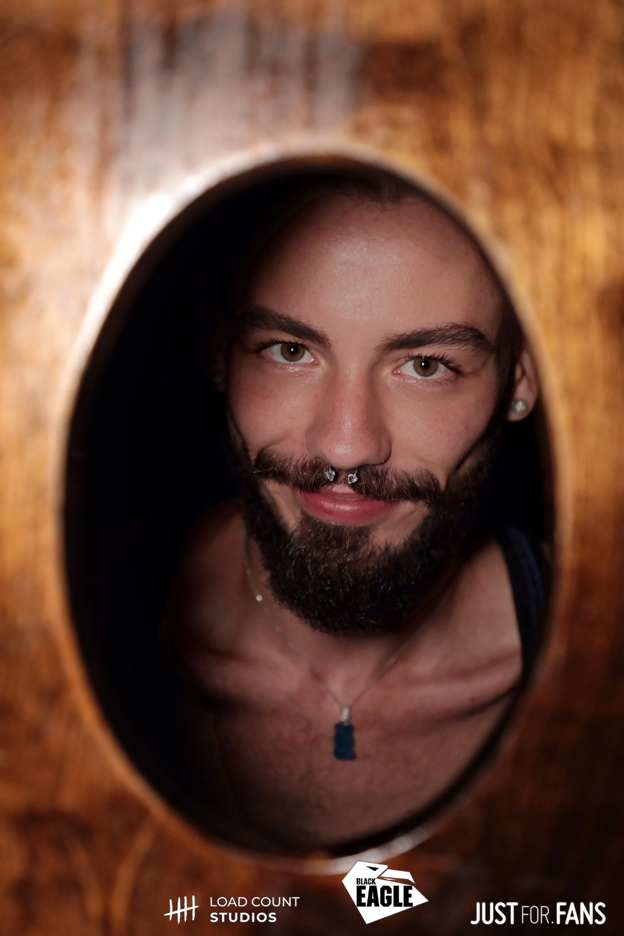 Person with beard and septum piercing smiling through a wooden frame.
