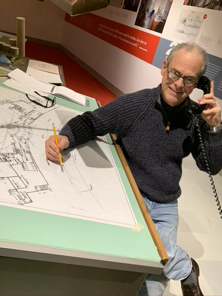 A man sitting at a table with architectural blueprints, talking on a landline phone, with a pencil in his hand, in a museum or exhibit space.