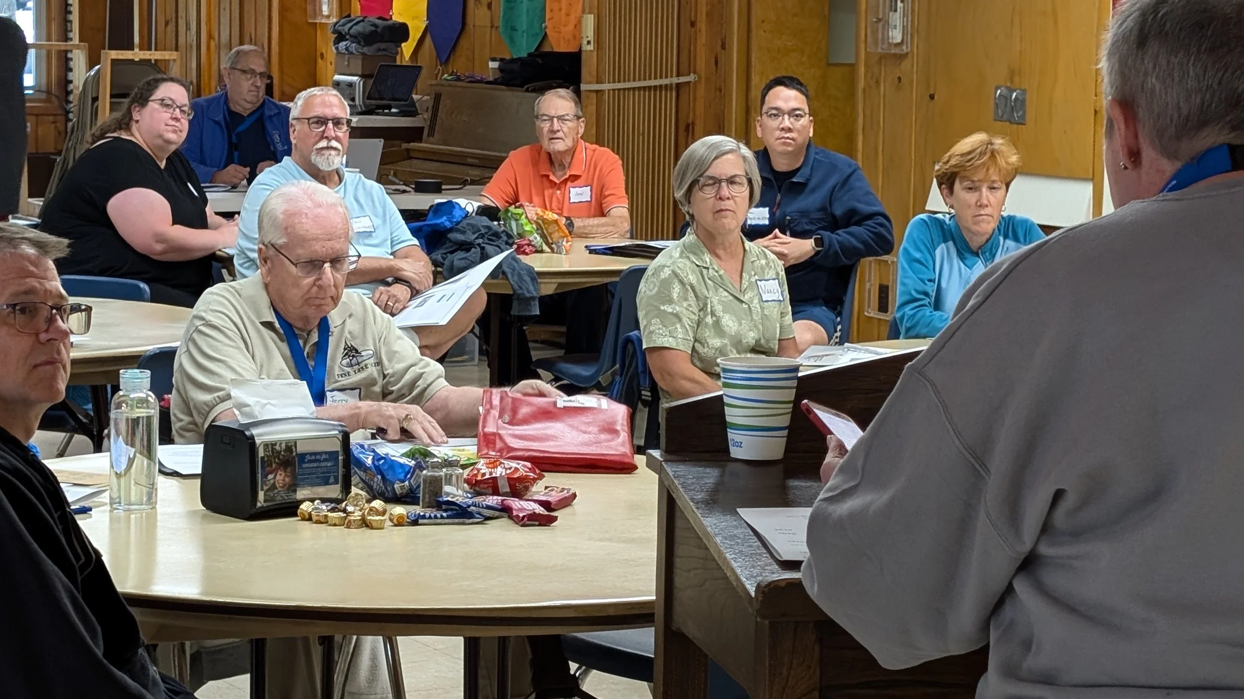 Group of people attending a meeting or class in a rustic wooden room, sitting at tables with papers, snacks, and drinks, listening to a speaker at the front.