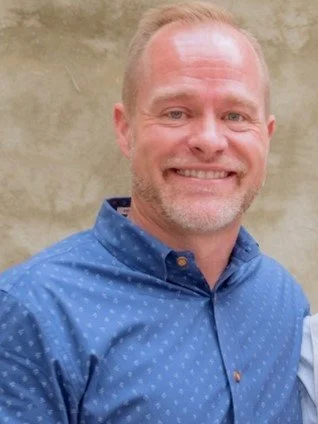 Portrait of a middle-aged man with short light hair and a beard, smiling, wearing a blue button-up shirt, standing outdoors against a beige wall.