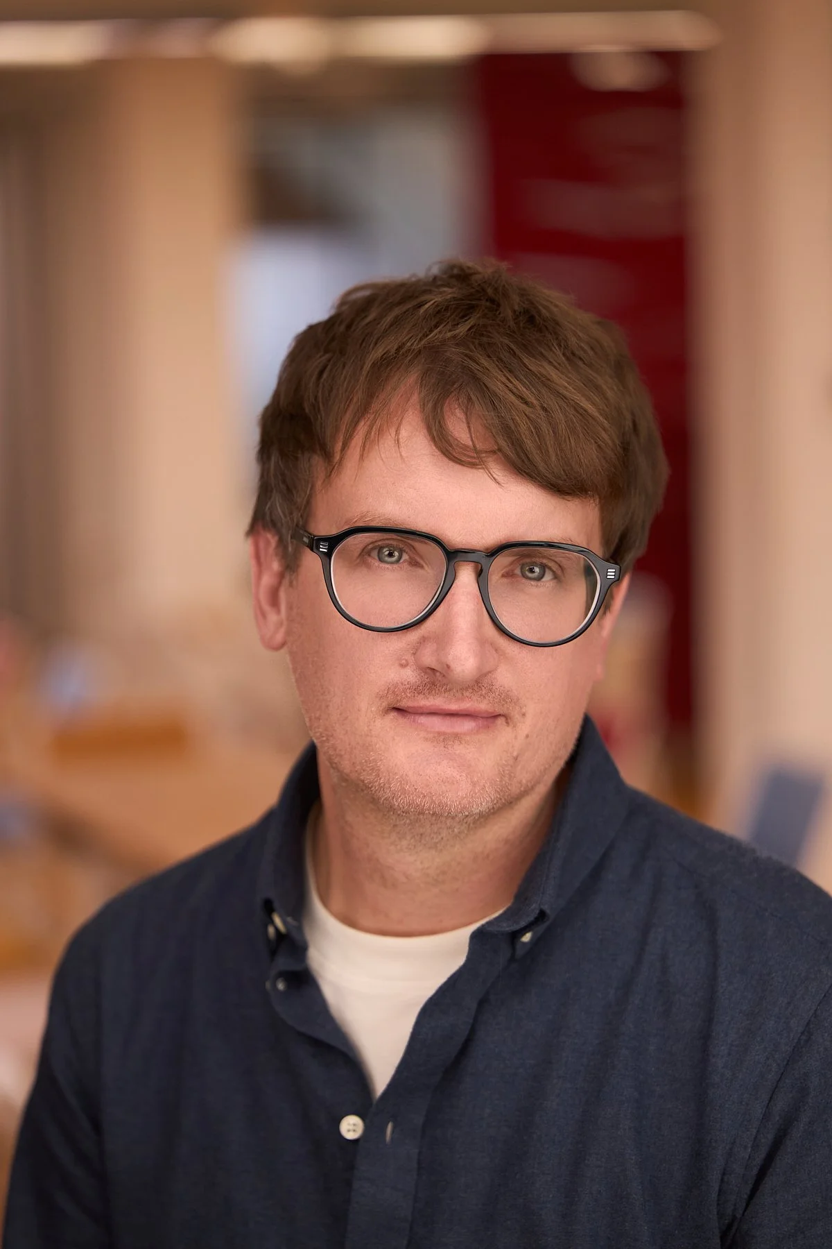 A man with brown hair and glasses looking at the camera, wearing a dark blue shirt with a white T-shirt underneath, in an indoor setting.