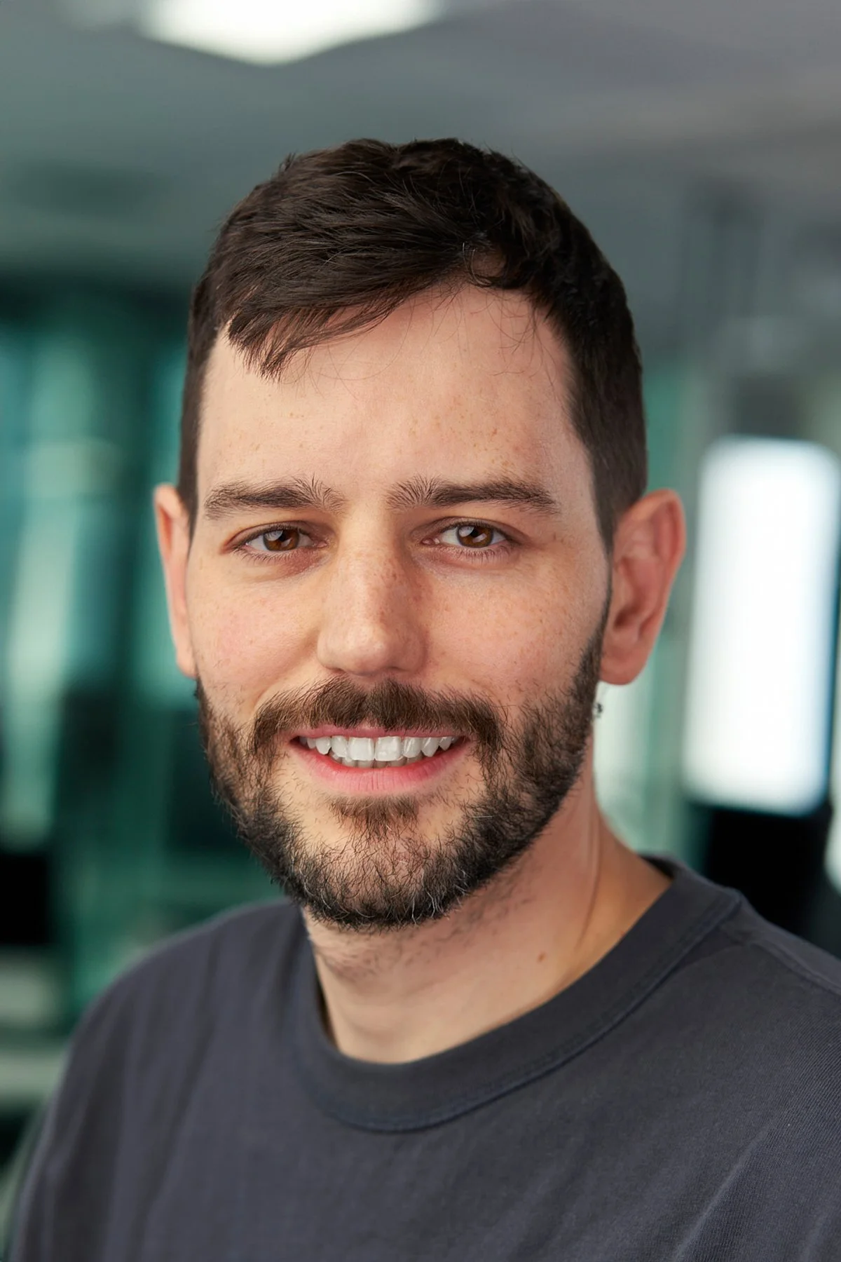 A young man with brown hair and a beard, smiling, wearing a black t-shirt, in an indoor setting with a blurred background.