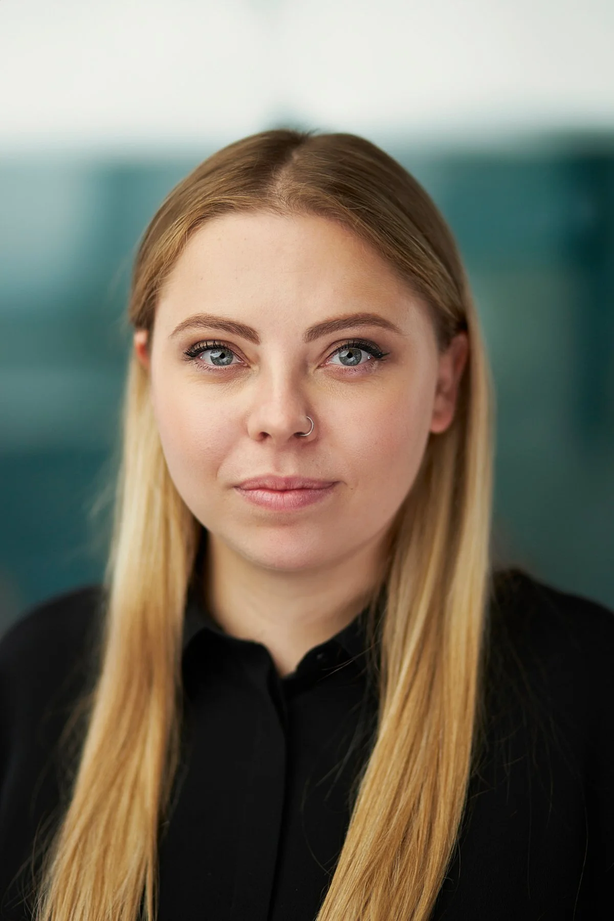 Close-up portrait of a young woman with long blonde hair, blue eyes, wearing a black shirt and a nose ring, with a blurred background.