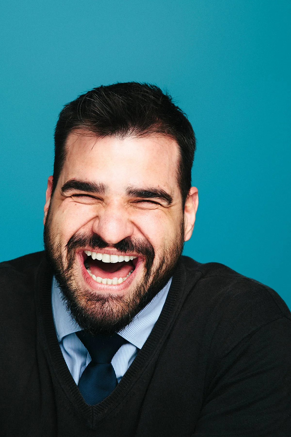 A man with dark hair and a beard laughing with his mouth wide open, wearing a black sweater, light blue collared shirt, navy tie, and standing against a blue background.