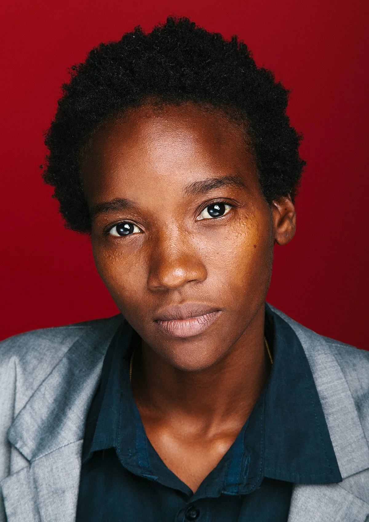 Close-up portrait of a young Black woman with short, natural curly hair, wearing a gray blazer and a dark shirt against a red background.