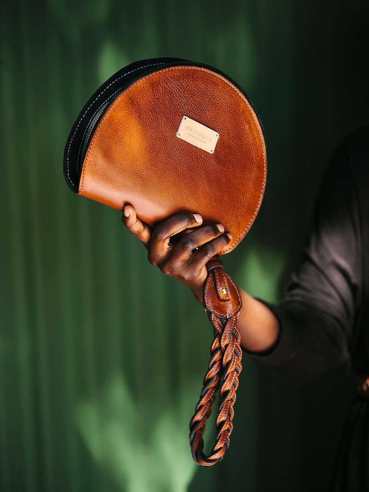 A person holding a semi-circular, brown leather handbag with a braided strap, against a dark green background.