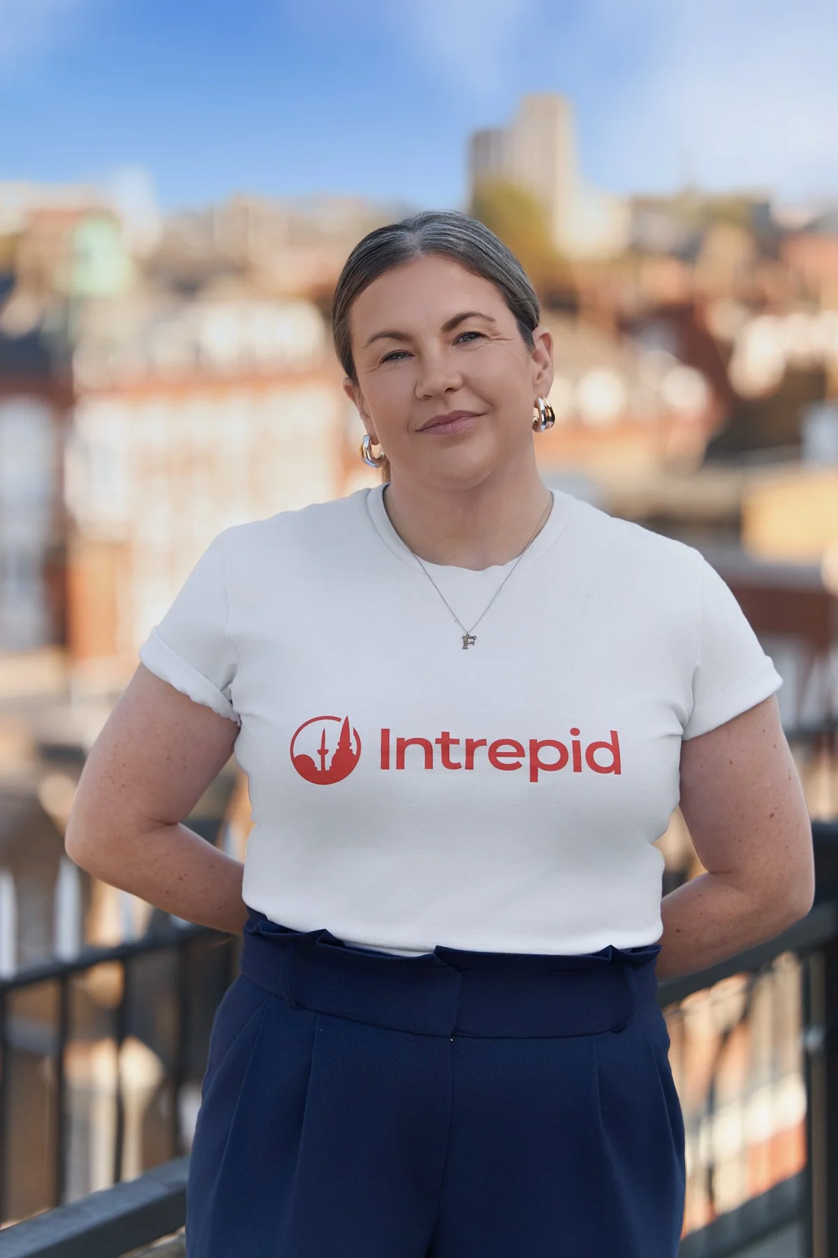 A woman outdoors in front of a cityscape, wearing a white T-shirt with the red 'Intrepid' logo and navy pants, smiling at the camera.