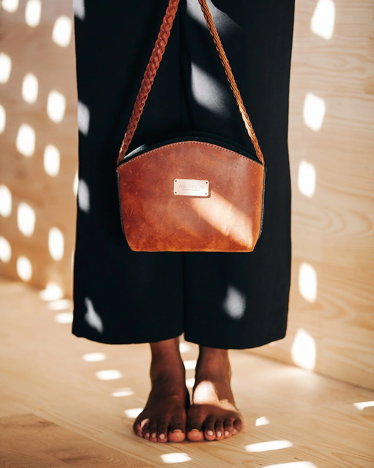 A person standing barefoot on a wooden floor, wearing black pants, with a brown leather crossbody bag hanging from their shoulder.