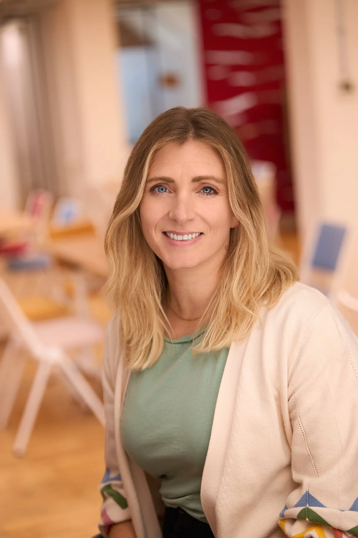 A smiling woman with blonde hair and blue eyes wearing a green top and a beige cardigan, sitting in a room with blurred background that includes chairs and tables.