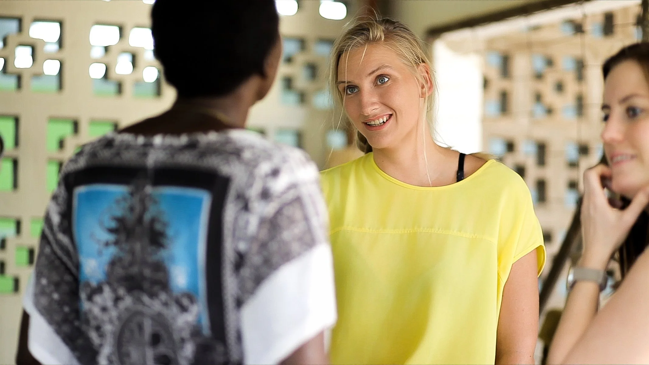 A group of young women and a young man having a conversation outside near a building with a patterned wall. The woman in the yellow shirt is smiling while talking to the man, who has his back to the camera. Another woman is visible on the right side,