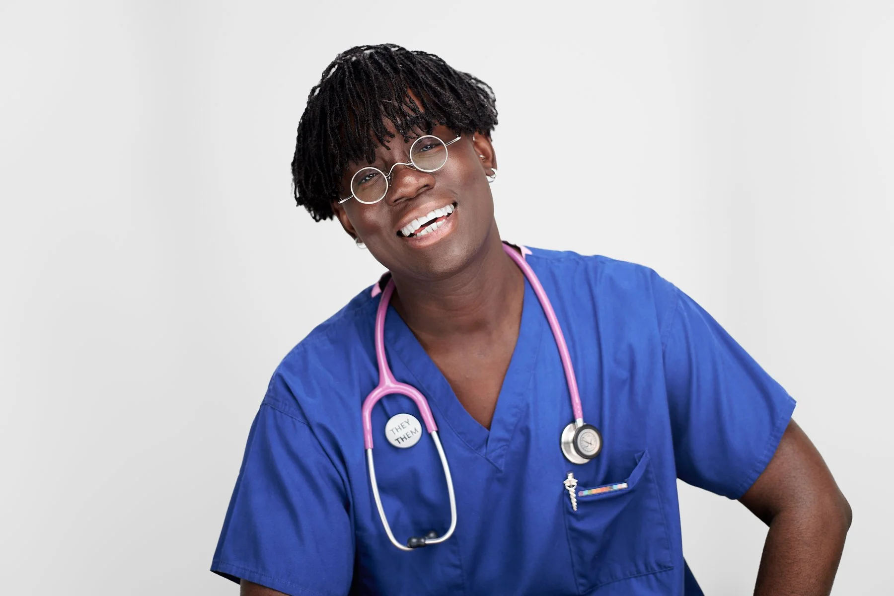 A smiling healthcare worker in blue scrubs with glasses and a stethoscope around their neck, standing against a plain white background.