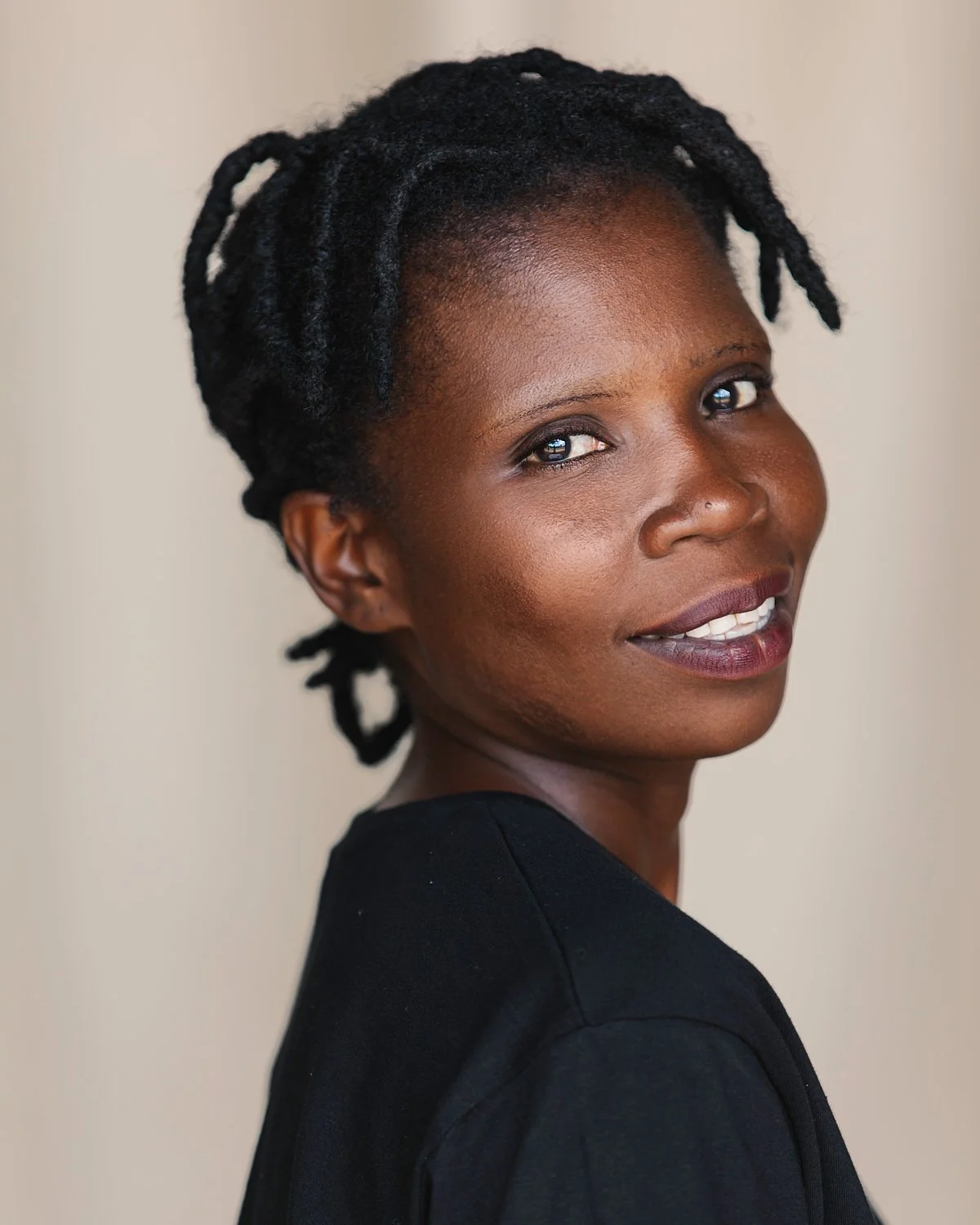 close-up portrait of an African American woman with short, dreadlocked hair, smiling softly, wearing a black top, against a neutral background