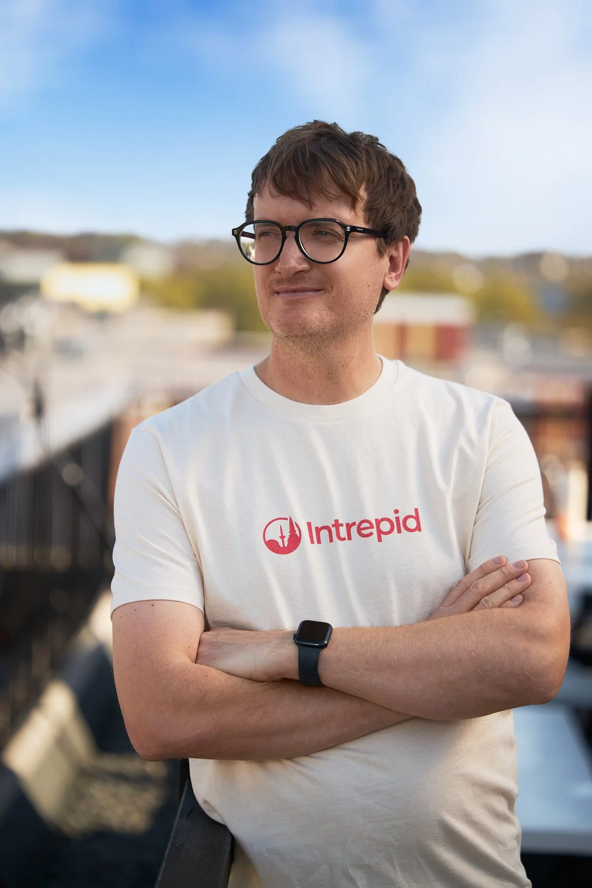 Man with brown hair and glasses wearing a white T-shirt with 'Intrepid' logo, standing outdoors with arms crossed, a blurred cityscape background, and a blue sky.