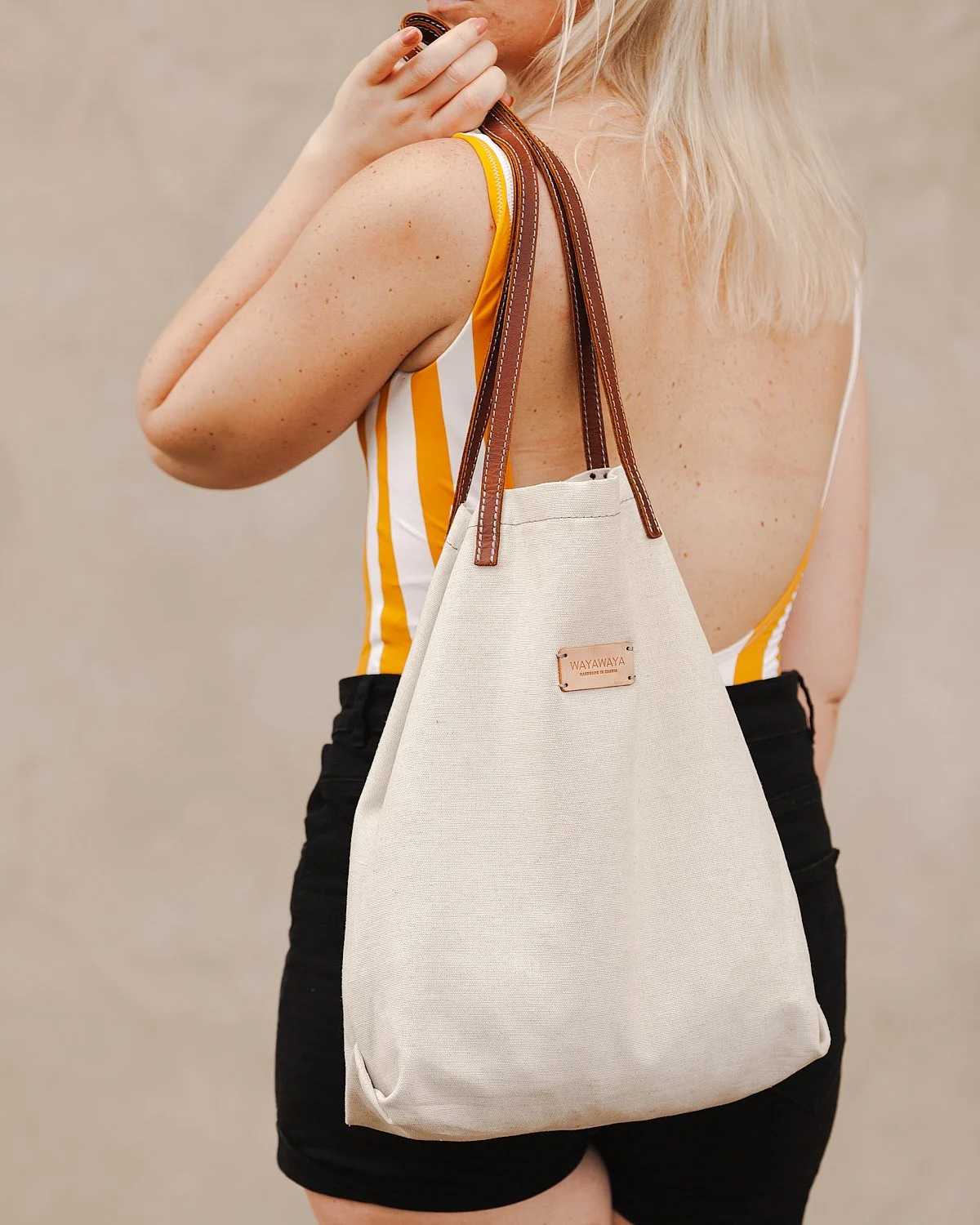 A woman with blonde hair holding a beige tote bag with brown leather straps over her shoulder, standing against a neutral background.